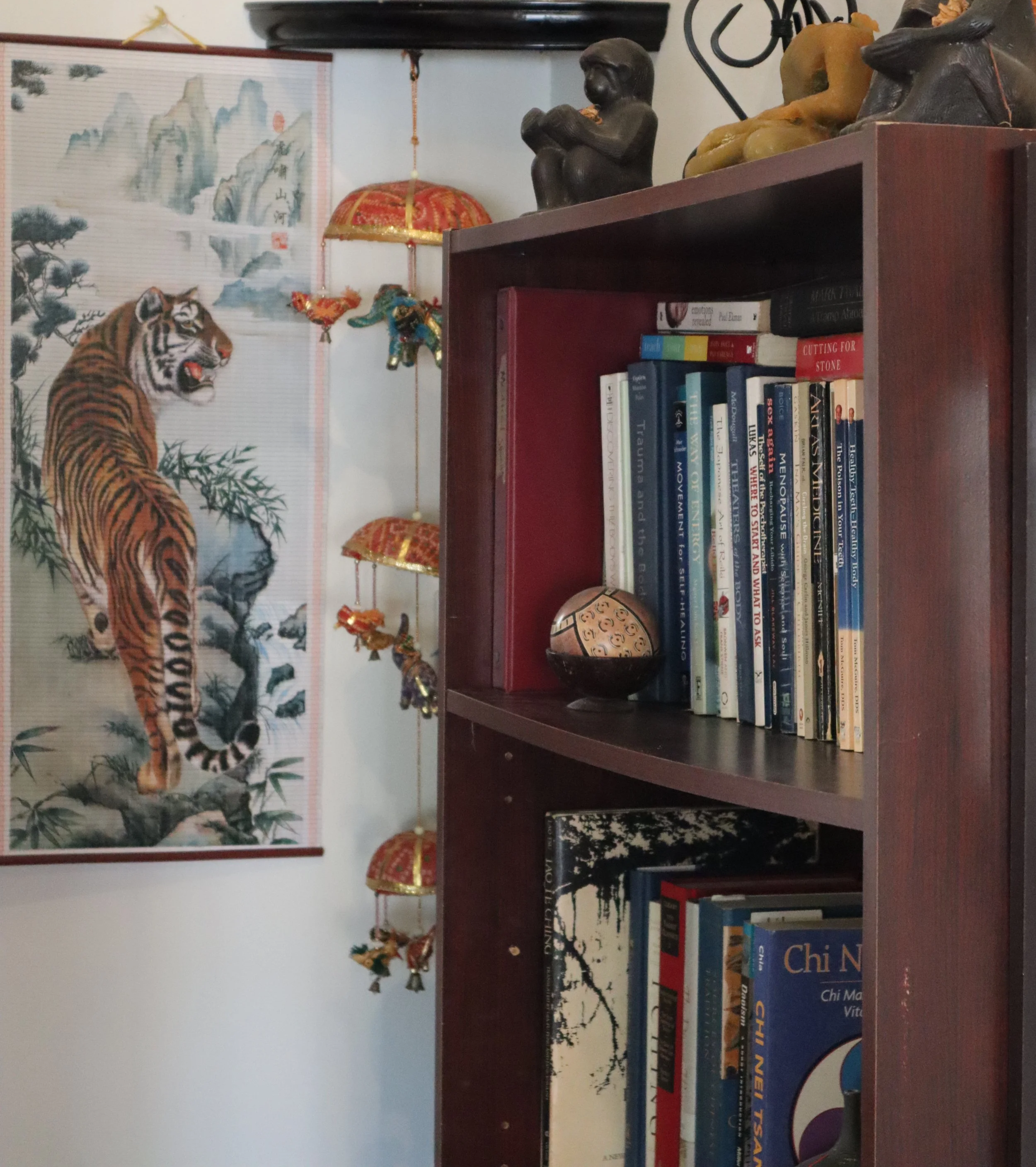 A bookshelf with a decorative Chinese scroll featuring a tiger and mountain landscape, along with Asian-style ornaments, a small bowl, and various books.