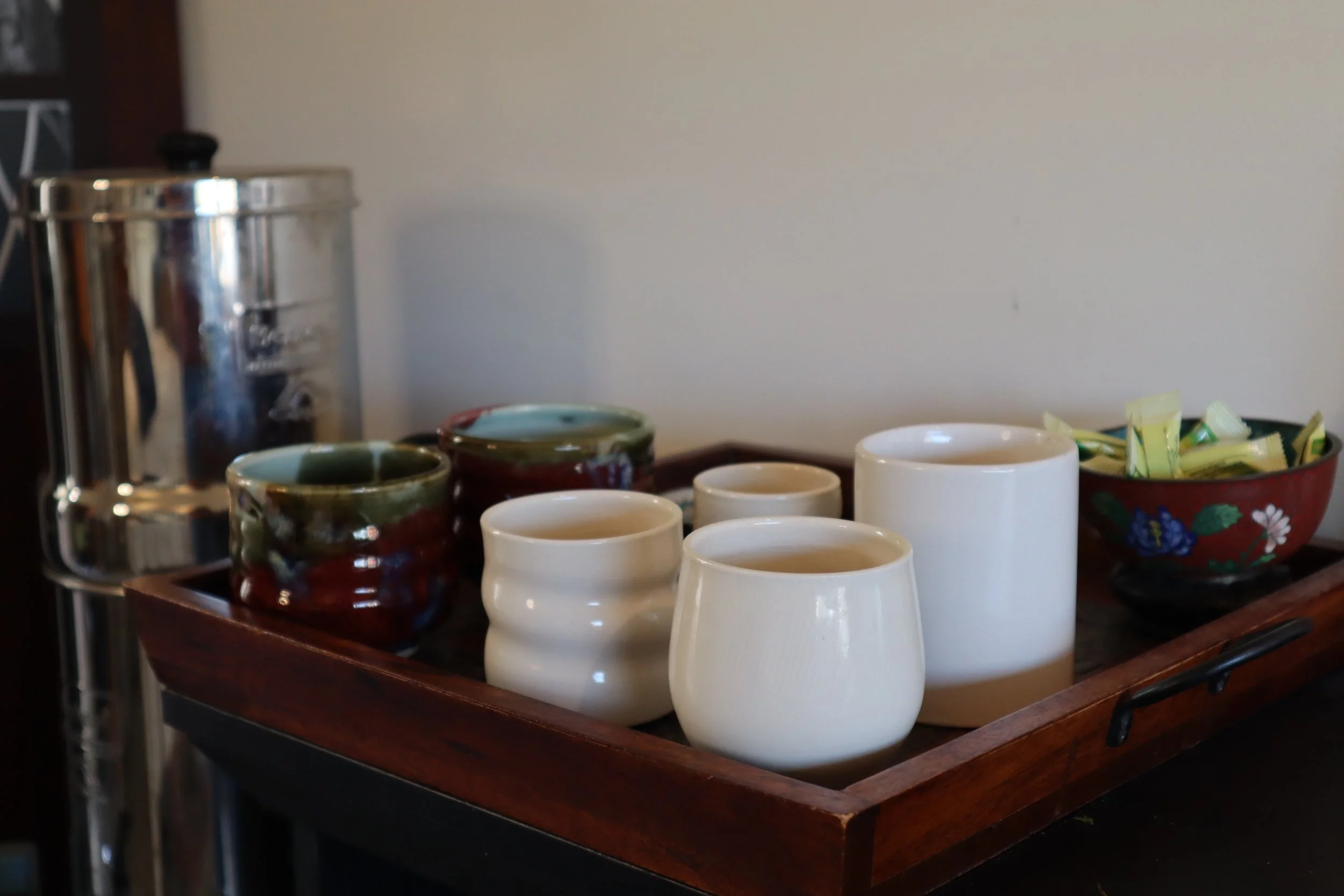 A wooden tray on a table holding several white cups, two small colorful bowls, and a larger floral bowl filled with wrapped sugar or sweetener packets, with a silver coffee urn and a plain white wall in the background.