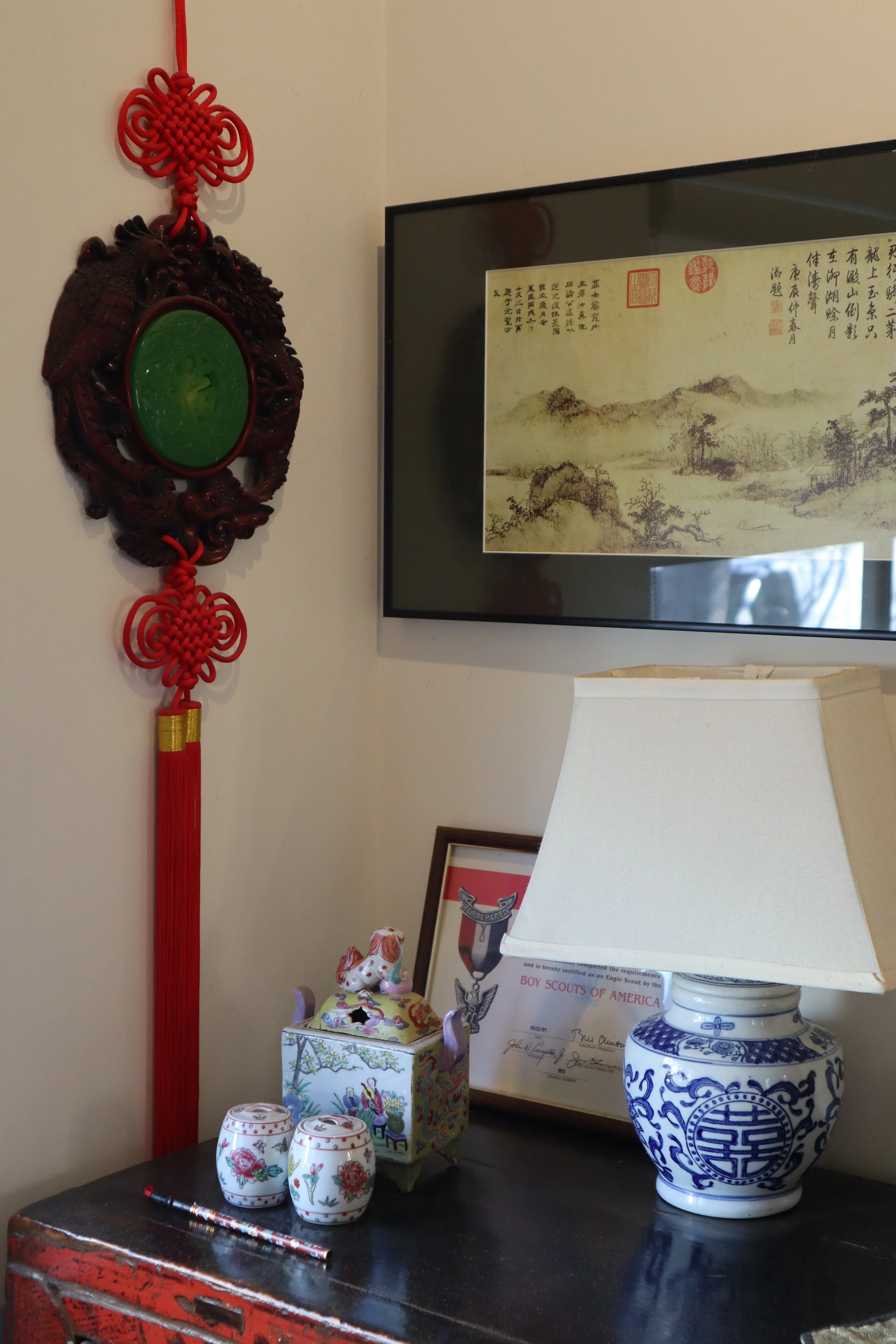 Decorative wall hanging, traditional Chinese style, with a red knot and green jade-like centerpiece, hanging beside framed Chinese calligraphy and artwork, on a wall above a dark wood table with porcelain and ceramic items, lamp, and framed certificate.