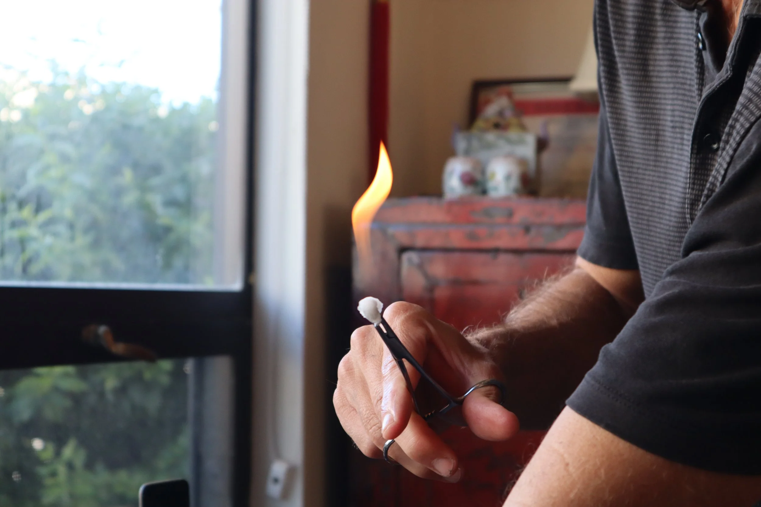 A person holding a lighter with a flame in front of a window, with various items on a shelf in the background.