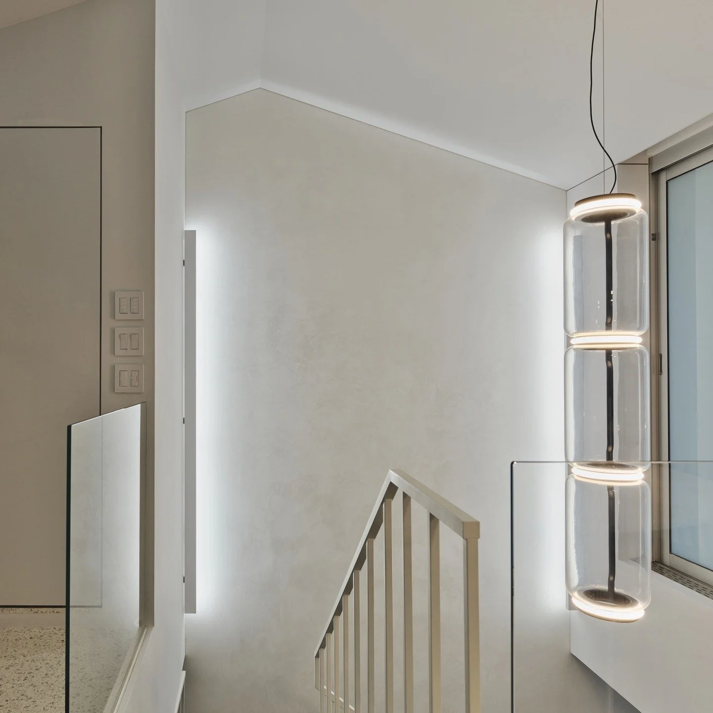 …
To connect the upper and lower floors, we installed a staircase. The design approach involved expanding and opening up this vertical space, complemented by large floor-to-ceiling windows that let natural light flood in. As a result, it has become the most dynamic feature of the project. However, since additional lighting is needed at night, we decided to select a pendant light to enhance the space.

Given that this is a tall, vertical space and the pendant is positioned right next to the dining table—a spot that catches the eye when ascending the stairs—it serves as a clear visual focal point. Therefore, the pendant needed to be a vertical, elongated fixture that could be viewed directly. With these requirements in mind, we quickly settled on the Noctambule by Flos

“Noctambule” refers to those night owls who are rarely seen during the day but come alive with splendor once night falls. This is the designer’s 