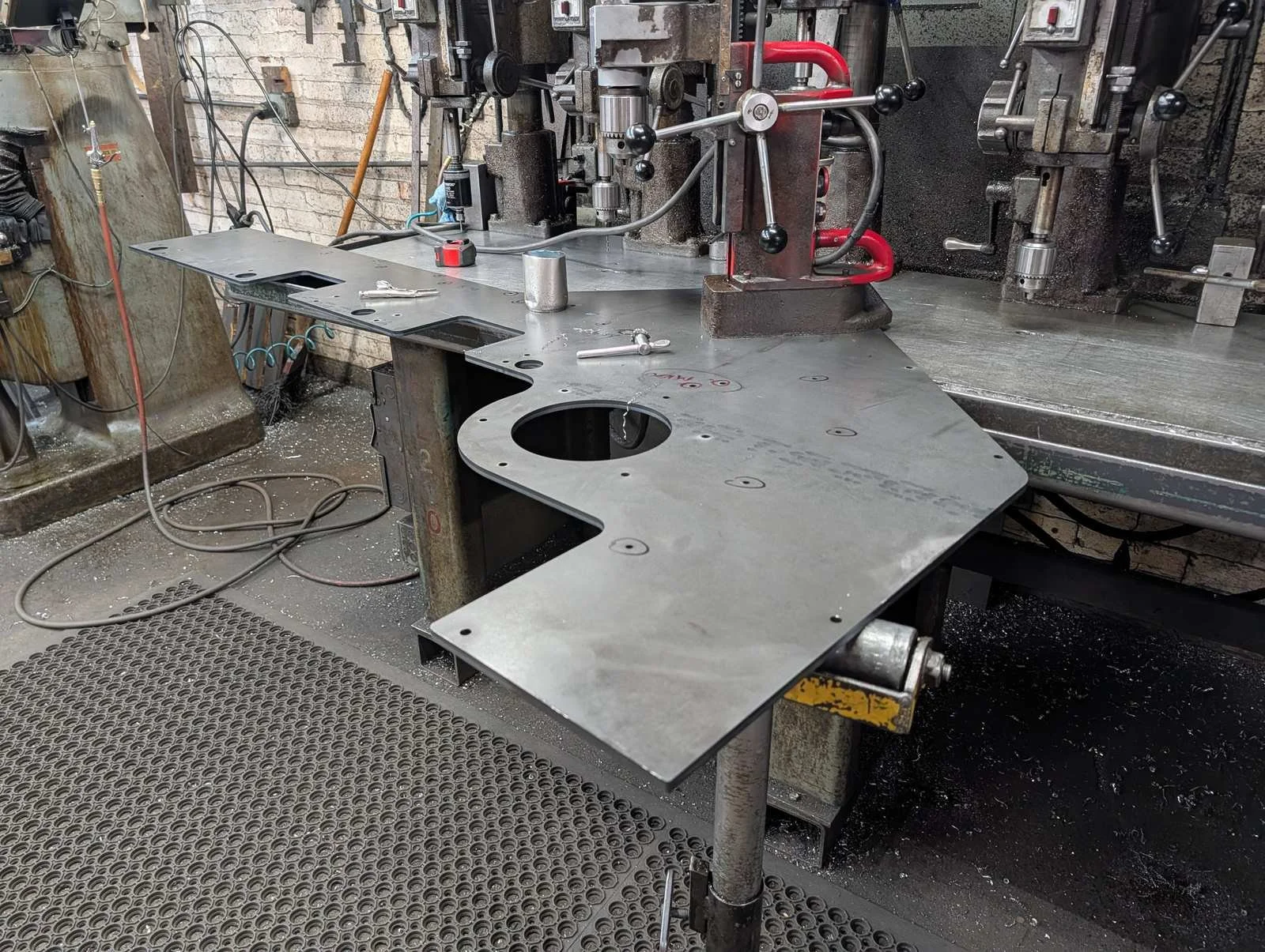 Large laser-cut steel plate with a round cutout and drilled holes at a drill press station