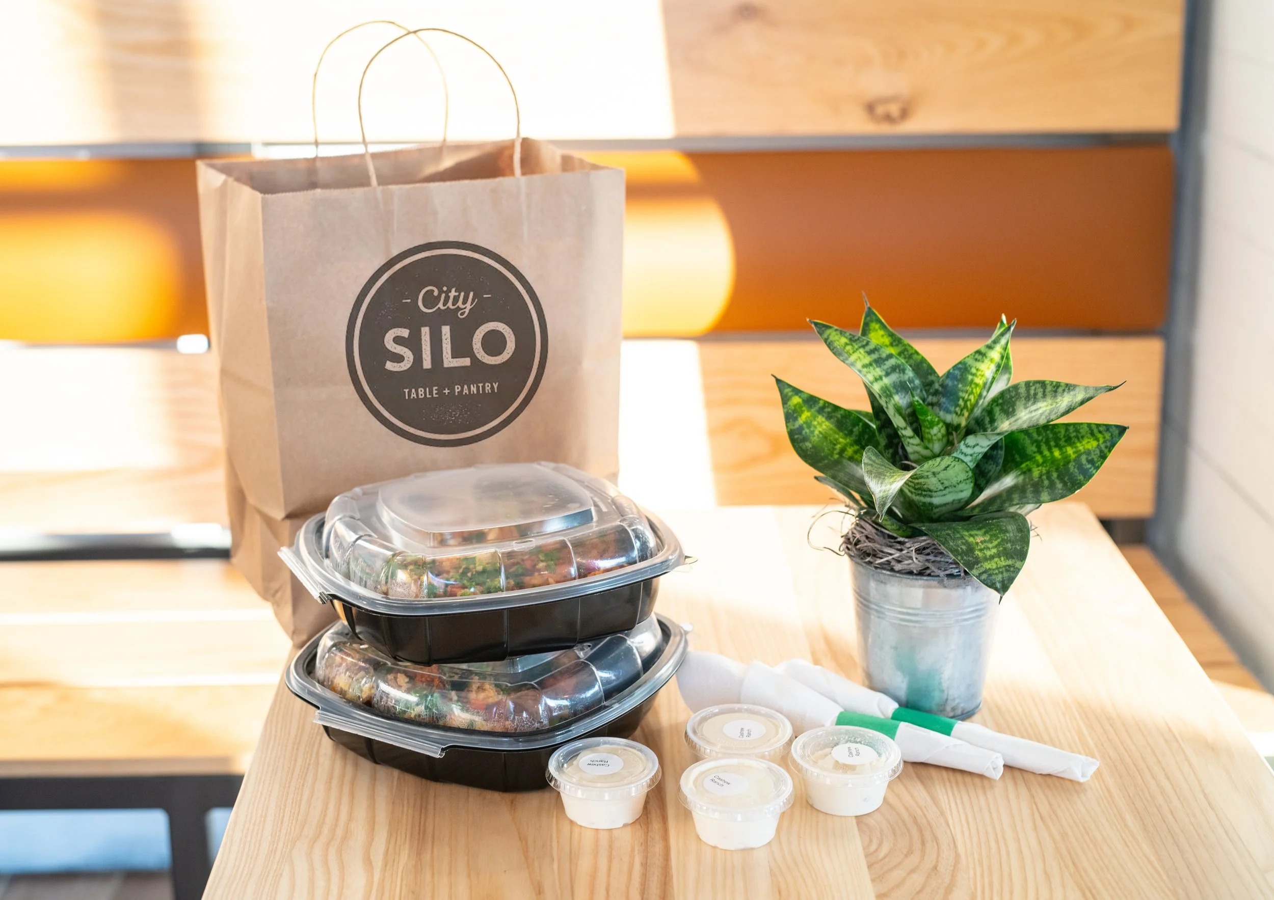 Food in to-go bags and containers sitting on a wooden table with a plant on the side