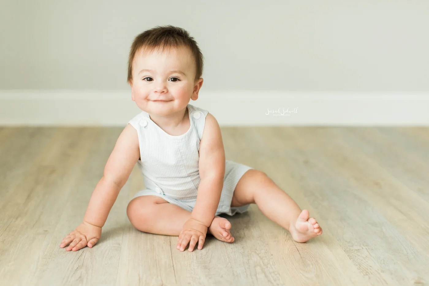 A baby boy sits on the floor and smiles.