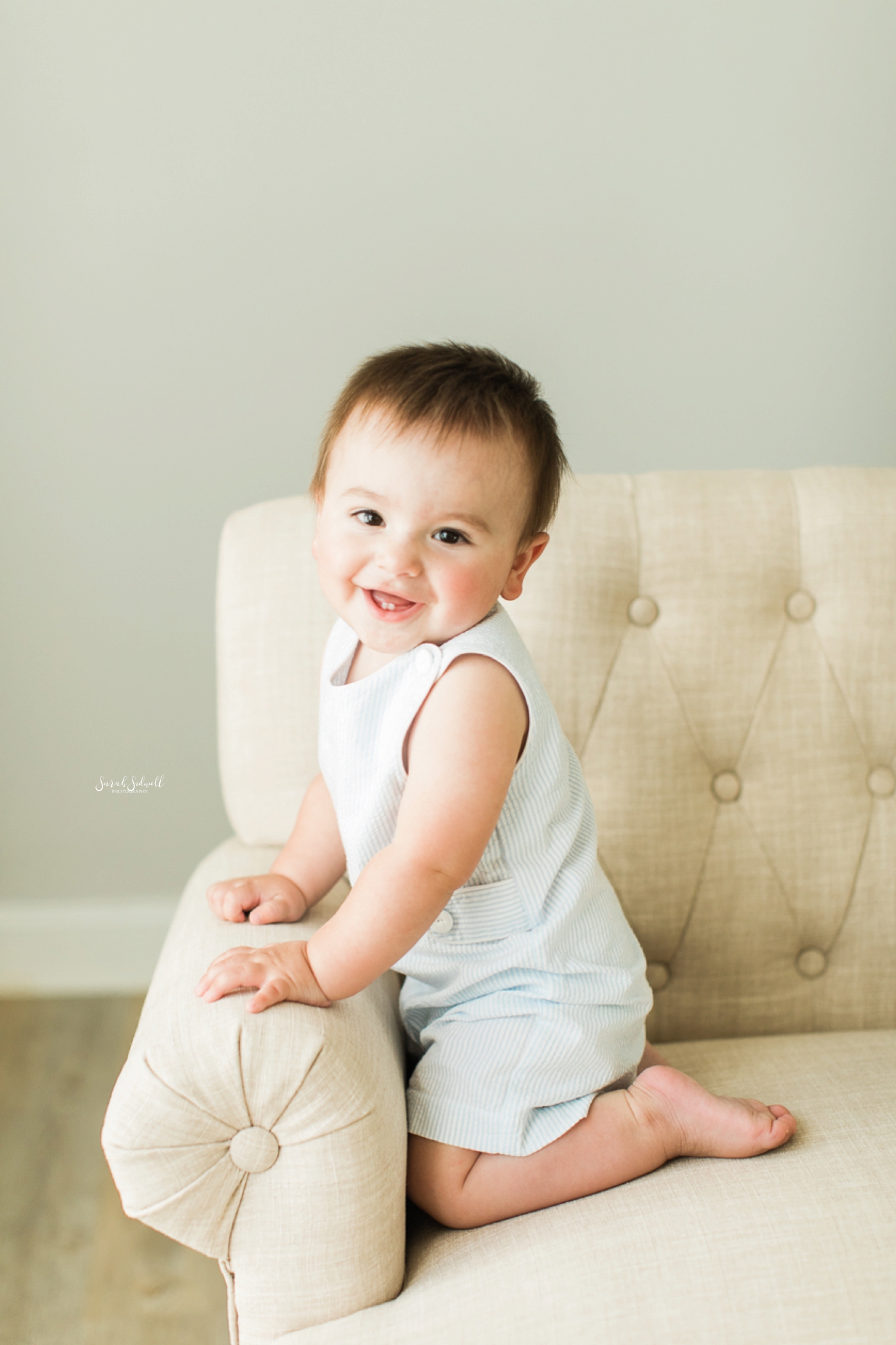 A baby boy sits on a white chair during family photography session.