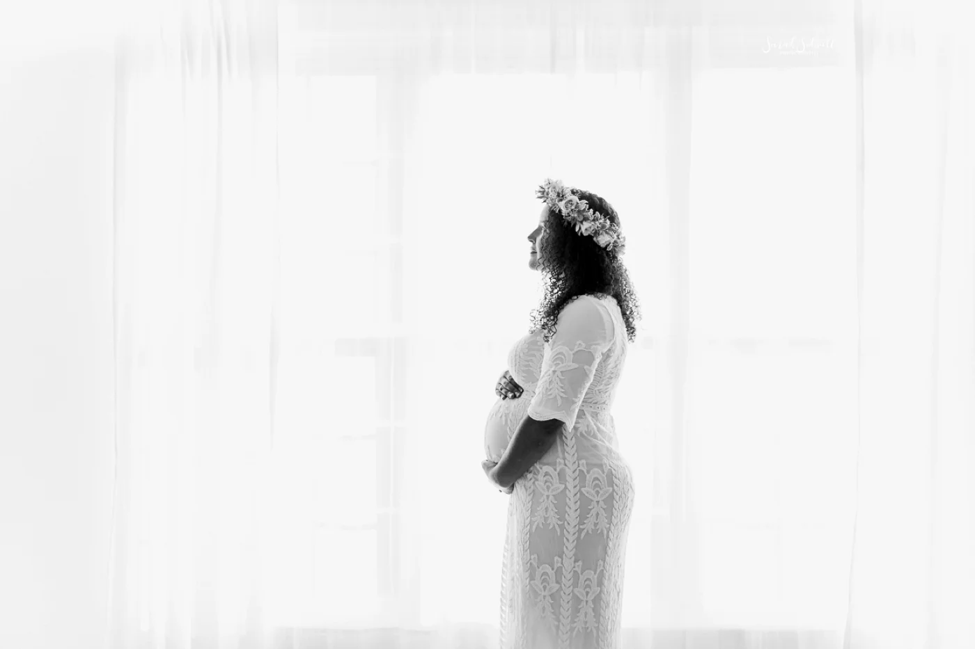 A pregnant woman in white stands by a window.