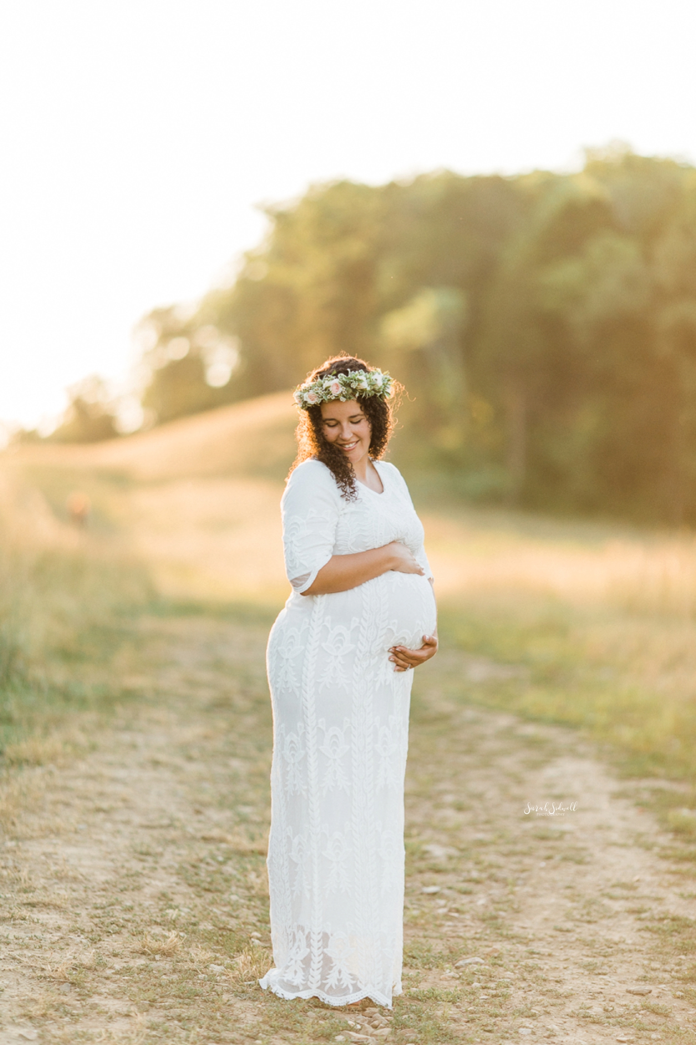 A pregnant woman wearing white and flower crown holds her belly.