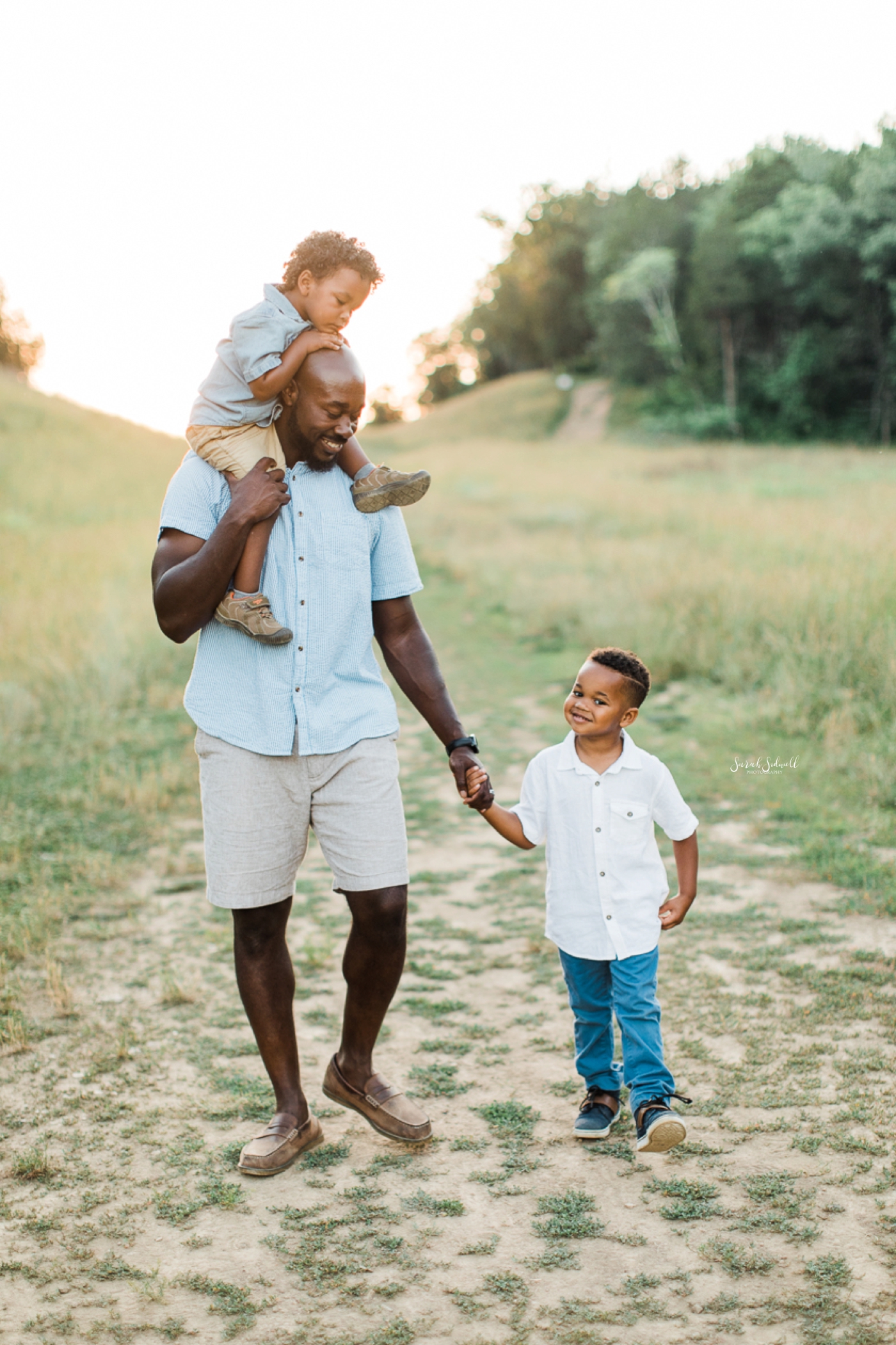 A man carries one son on his shoulders while holding the hand of his other son.