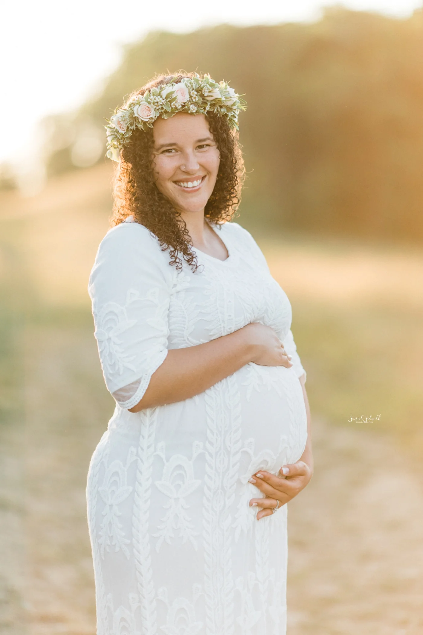 The sunset illuminates a pregnant woman wearing a white dress and flower crown.