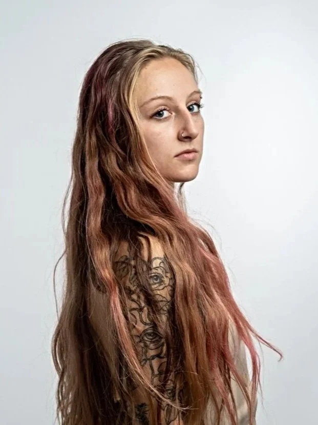 A young woman with long, wavy, reddish-brown hair, wearing a sleeveless top with tattooed arms, standing against a plain light gray background.