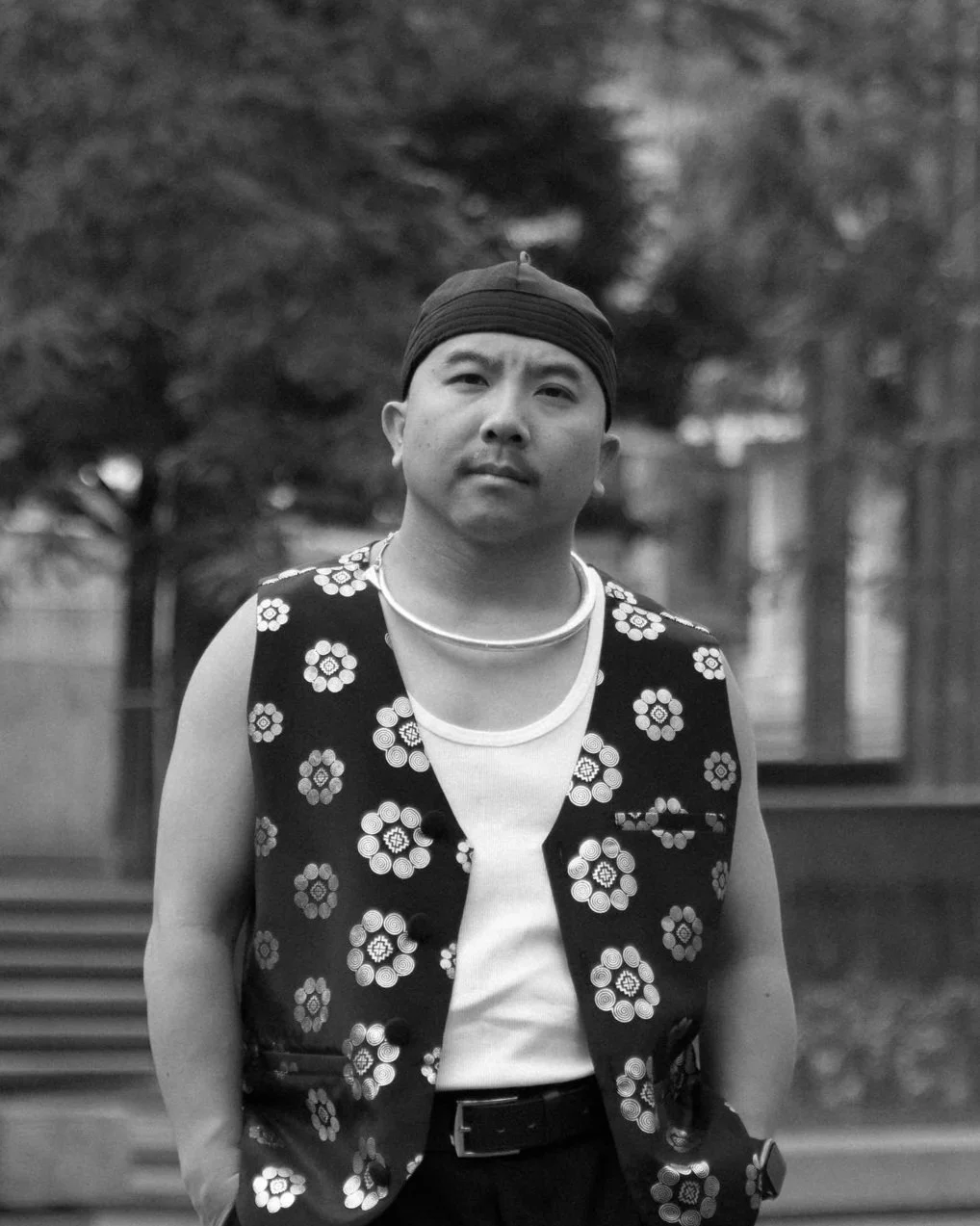 A person standing outdoors with trees in the background, wearing a patterned vest, a beaded necklace, and a head covering, captured in black and white.