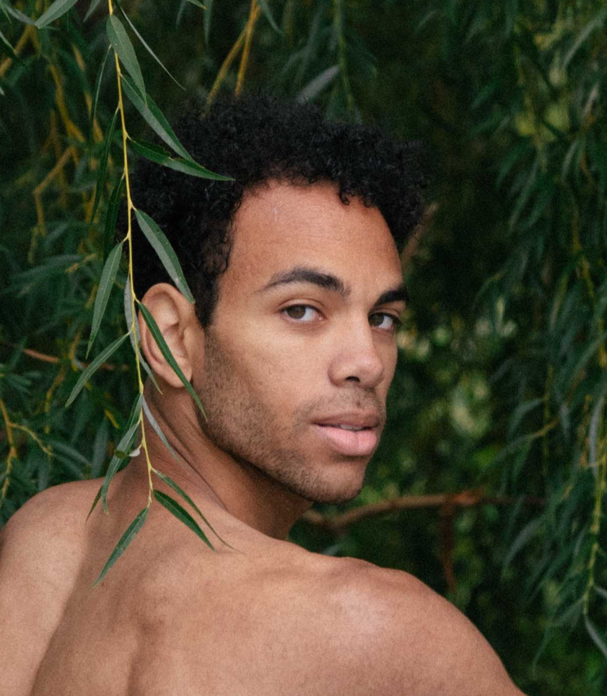 A shirtless man with dark curly hair and a light beard looking back over his shoulder among green leaves.