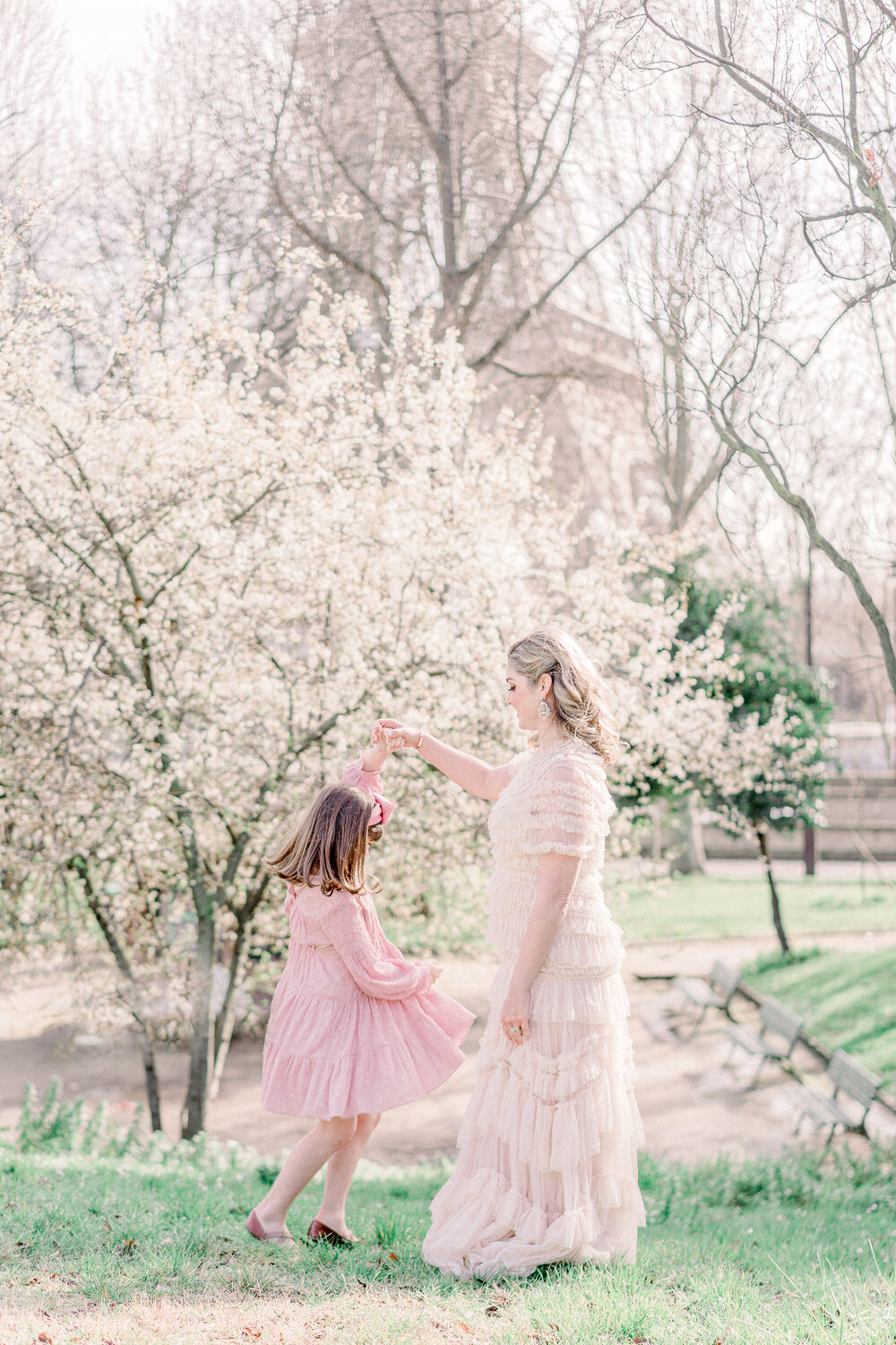  Mother Daughter dancing in grassy area in Paris Park. Mother is wearing a pink tiered tulle gown with a Blonde hair down do with defined eyes and liquid liner for Paris family photoshoot . Makeup and hair by english speaking makeup and Hair artist O