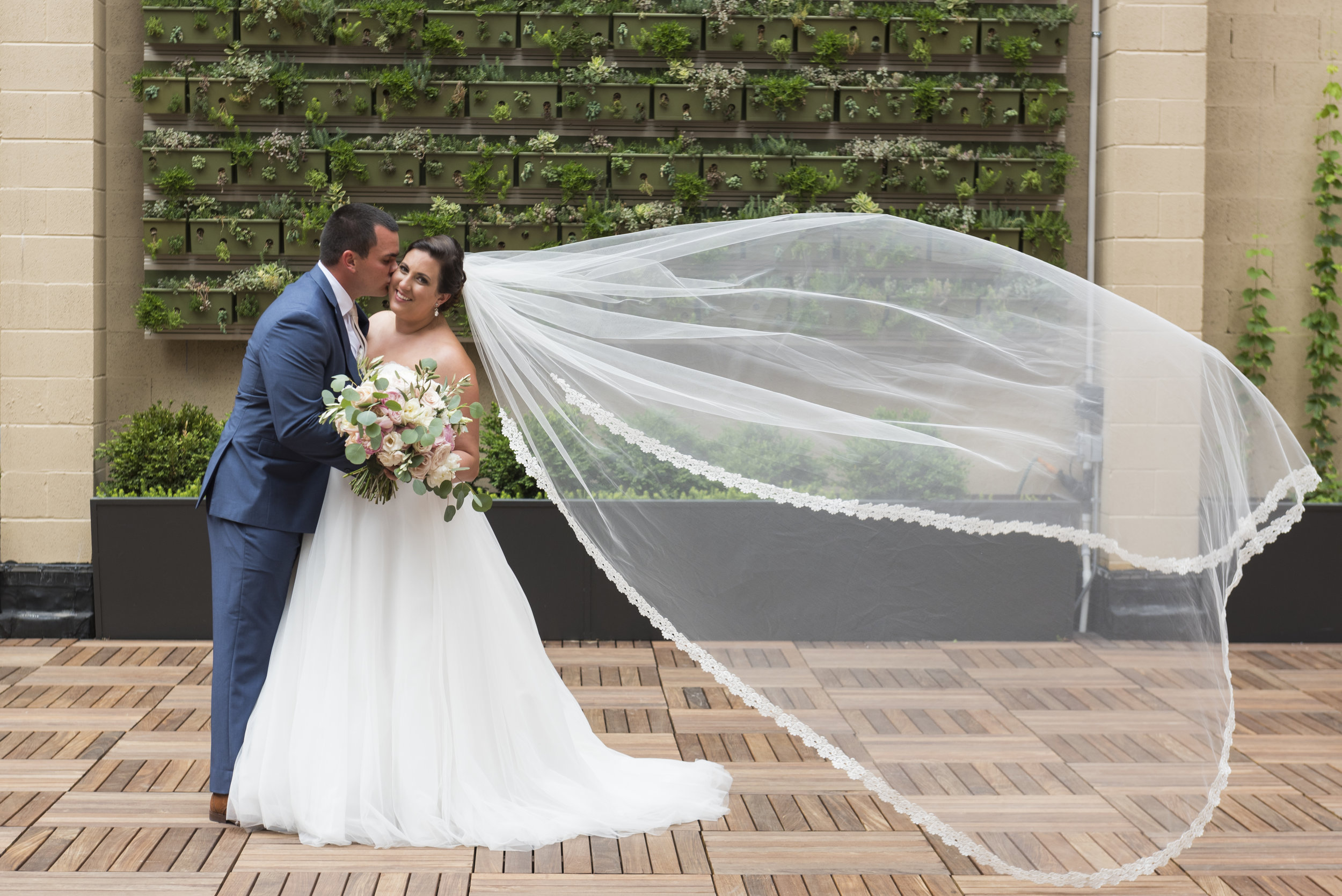 Groom and bride with floating veil outdoor portrait on Terrace in front of greenery succulent wall