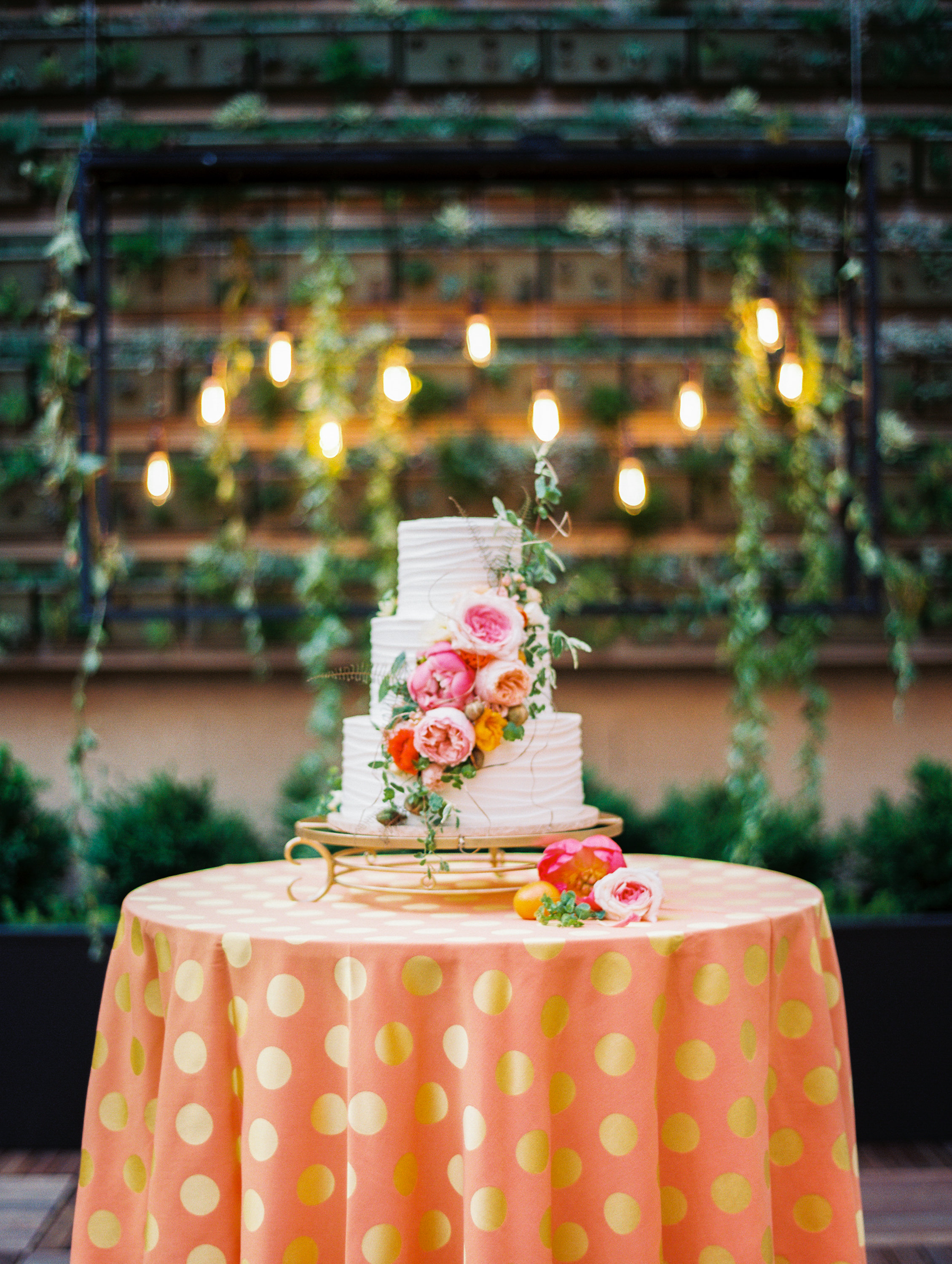 Wedding cake with roses and greenery in front of green wall with edison bulbs