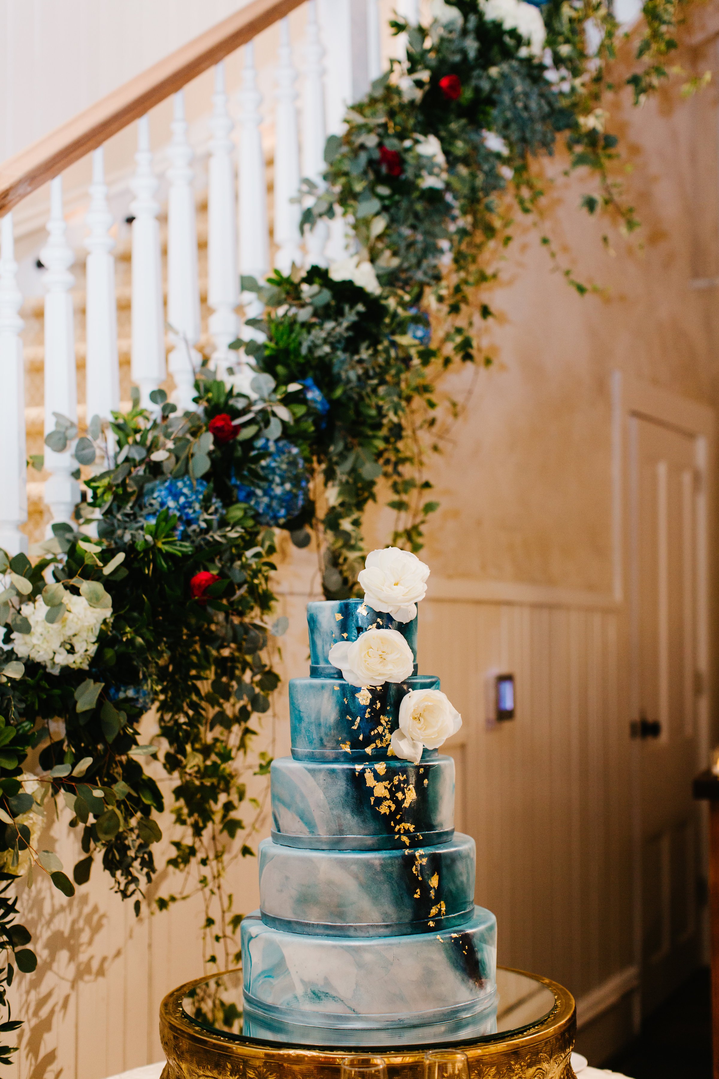 Blue &amp; gold wedding cake with white flowers next to a floral lined staircase