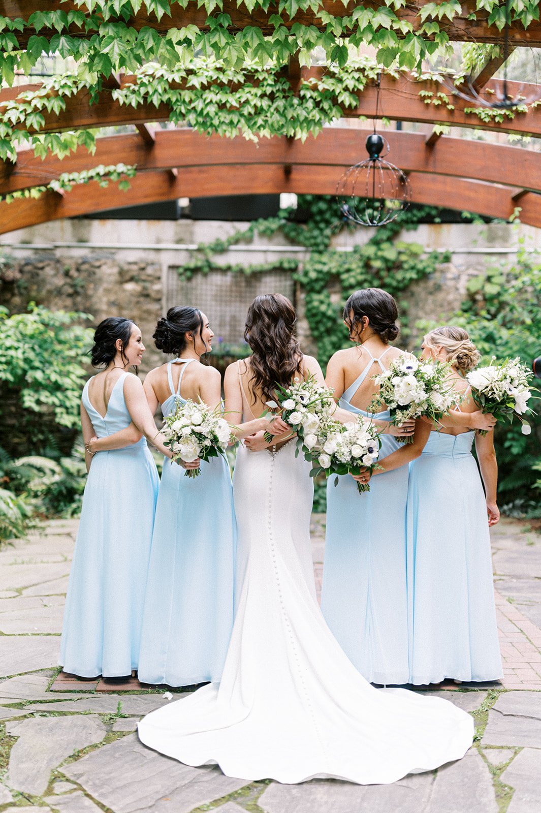 Bride and bridesmaids crossing arms across backs with white bouquets under greenery canopy