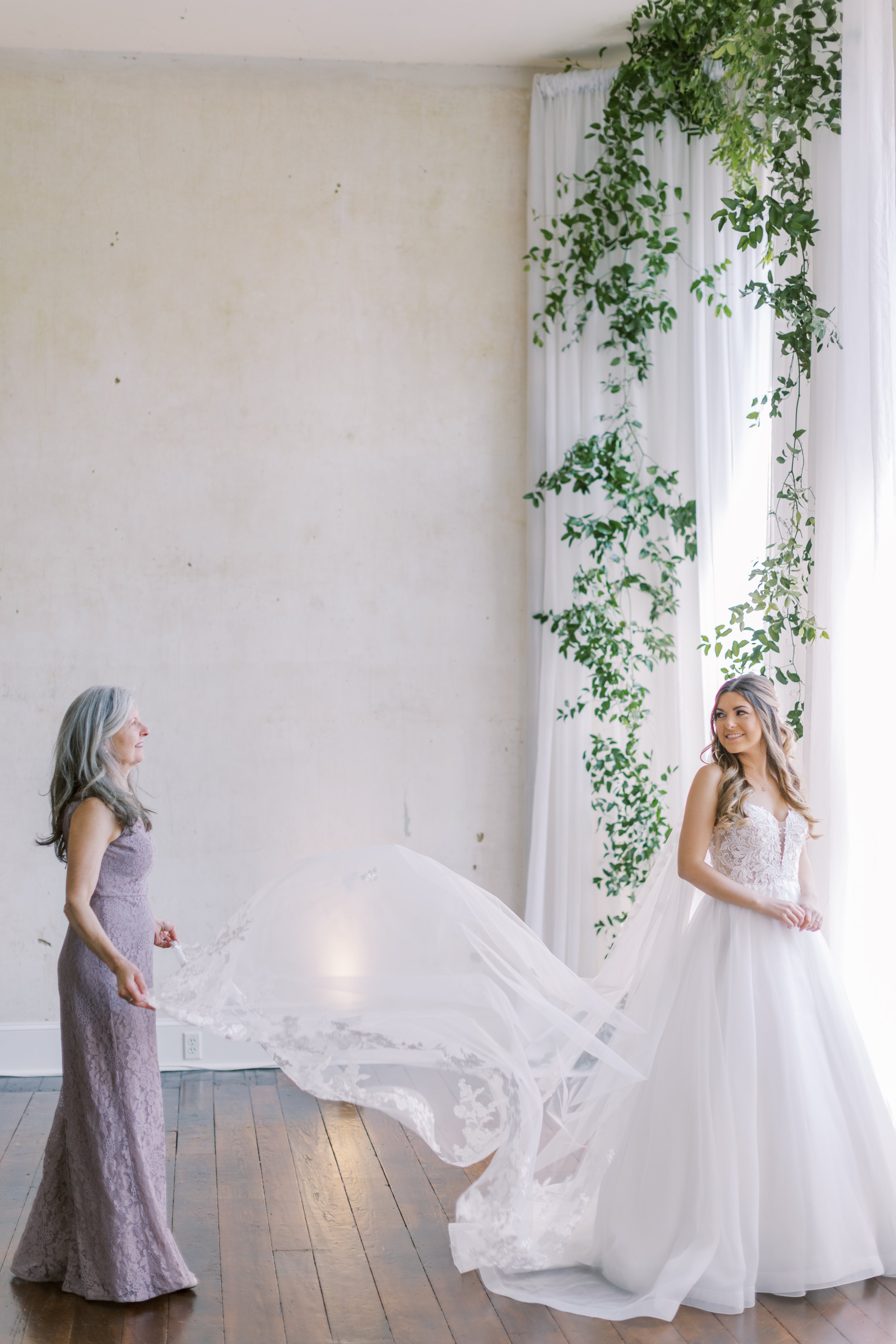 Mother of the bride helping daughter get ready in large room with greenery