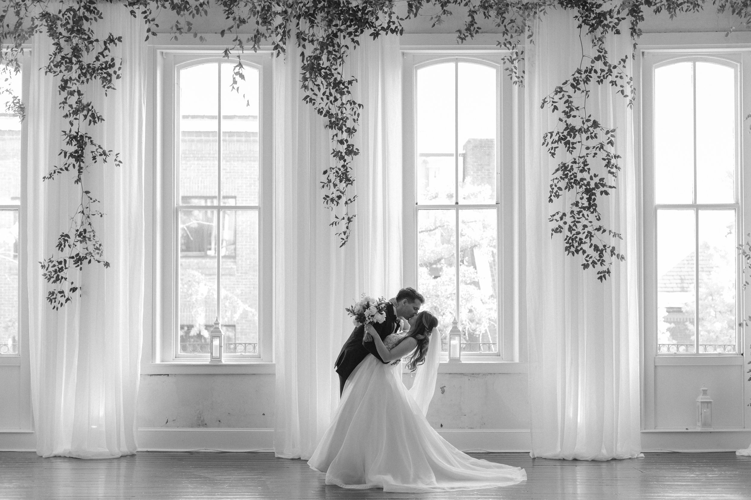 Bride and groom dramatic kiss in front of large windows with greenery hanging
