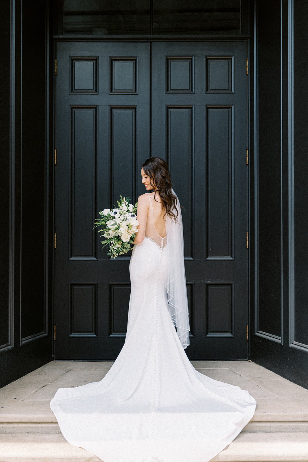 Classic bride with bouquet of white flowers looking at ground in front of black doors of Excelsior wedding venue