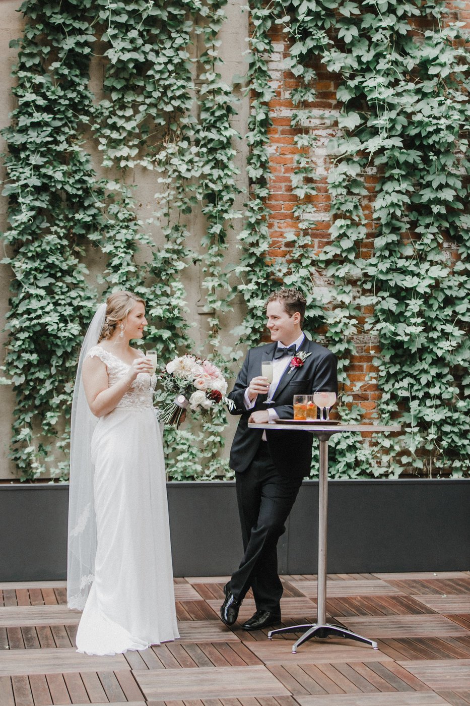 Bride and groom sipping cocktails in front of a green wall in the Garden Courtyard of Excelsior