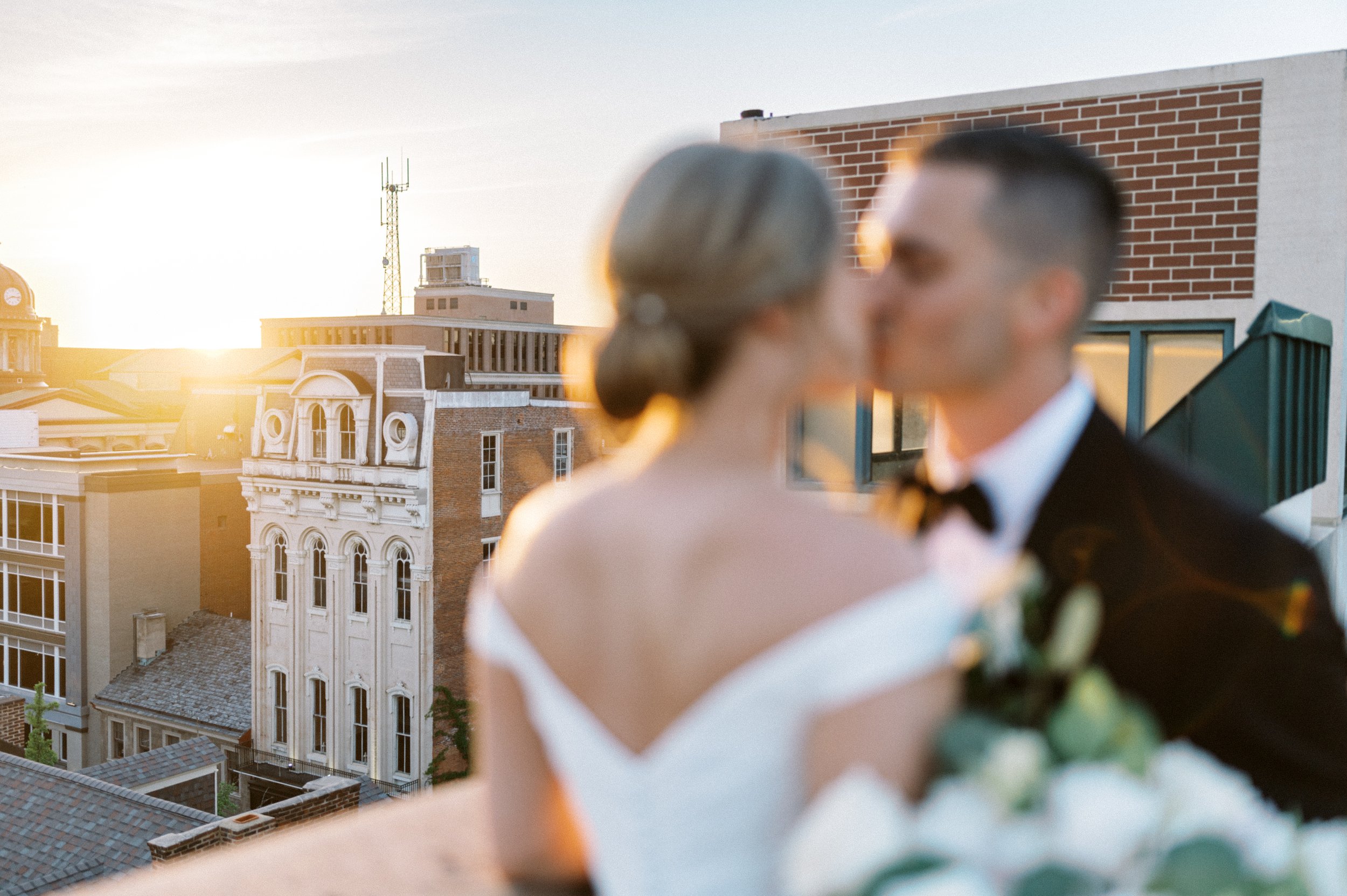 Bride and groom kissing out of focus in front of Excelsior exterior front facade at sunset