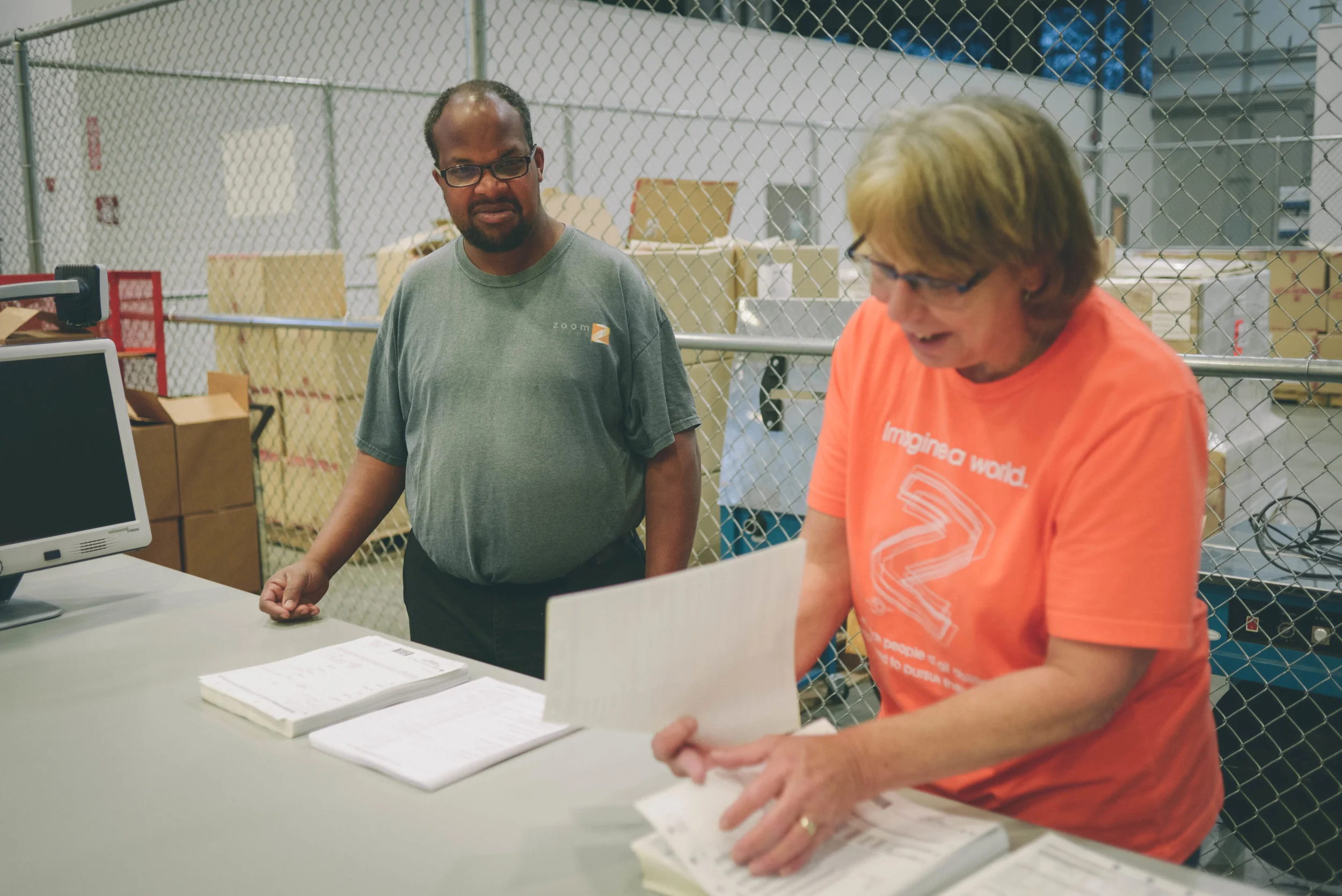 Image of two individuals in a warehouse setting. One looks on at the other sorting through some paper.