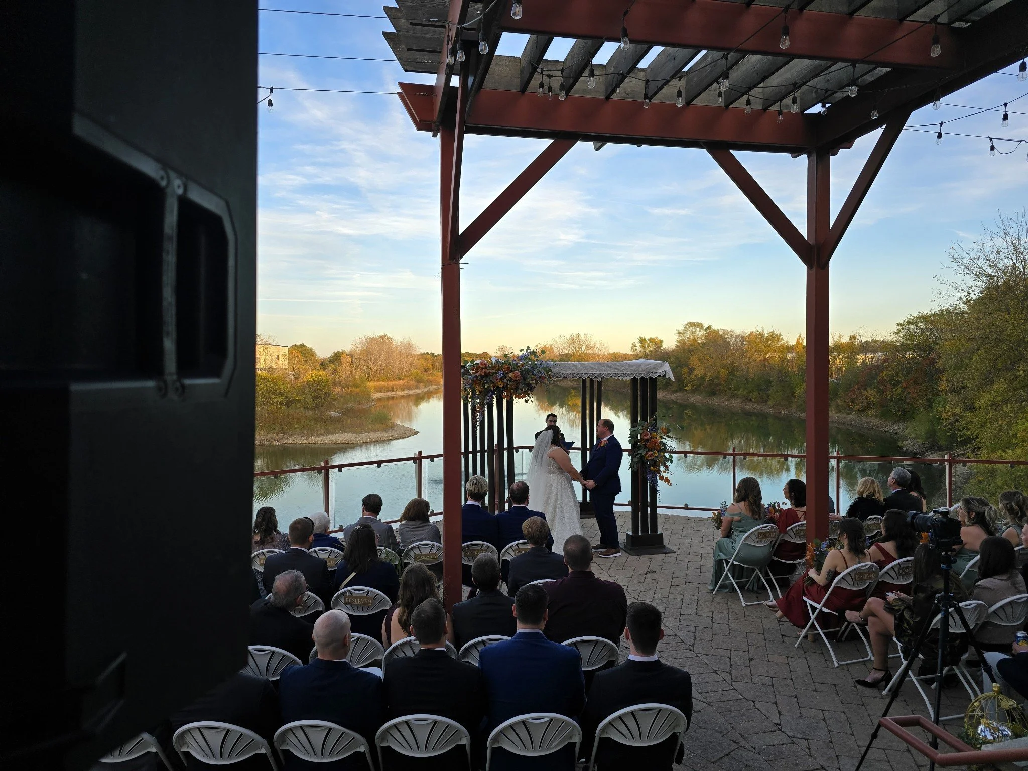 A couple getting married at an outdoor lakefront ceremony with guests seated on white chairs, officiant, and decorated arch, under a wooden pavilion at sunset.