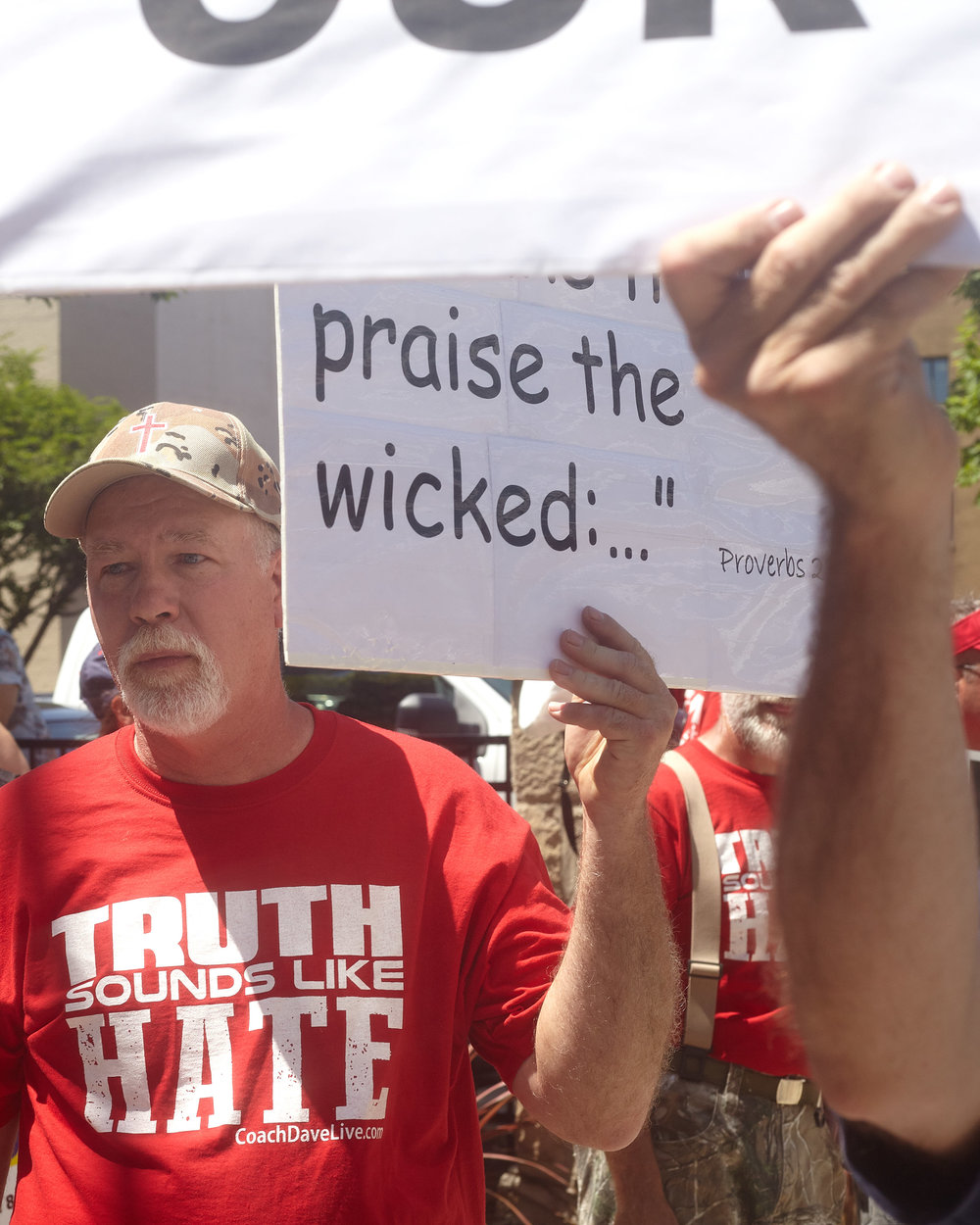 A counter protester wearing a shirt that states "Truth Sounds Like Hate" holds a sign with a biblical passage.