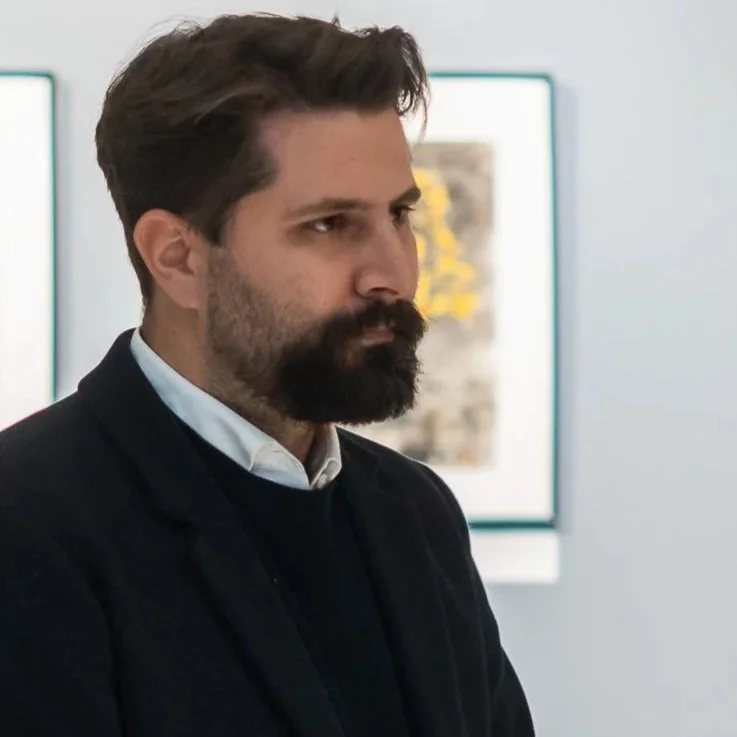 Artist Georgios Merianos standing in a gallery space with framed artworks visible in the background.