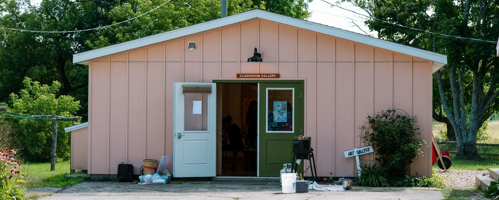 a small pink building labeled "Classroom Gallery" with double doors open, showing a glimpse of people inside. Art supplies and tools sit outside near the enterance, and a handmade "Art Gallery" sign points towards the building.