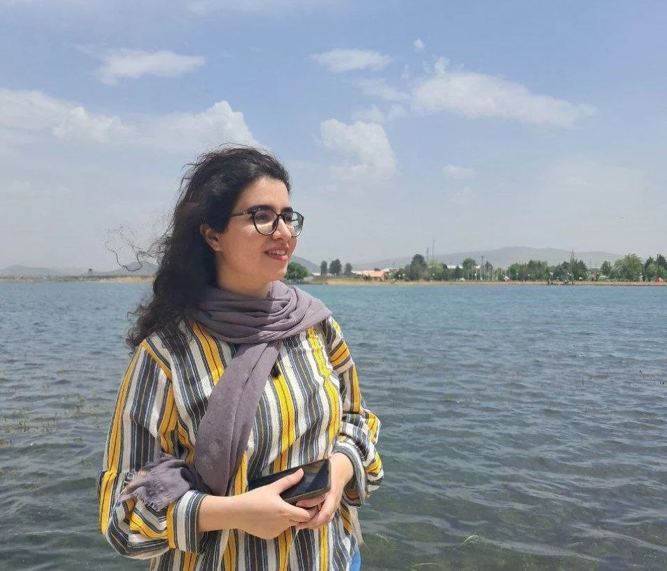 A young woman with long dark hair and dark glasses looks to off to the side. She is wearing a gray scarf over a yellow, white, and gray striped dress. In the background is a vast expanse of blue water, and the hint of buildings, trees, and a mountain