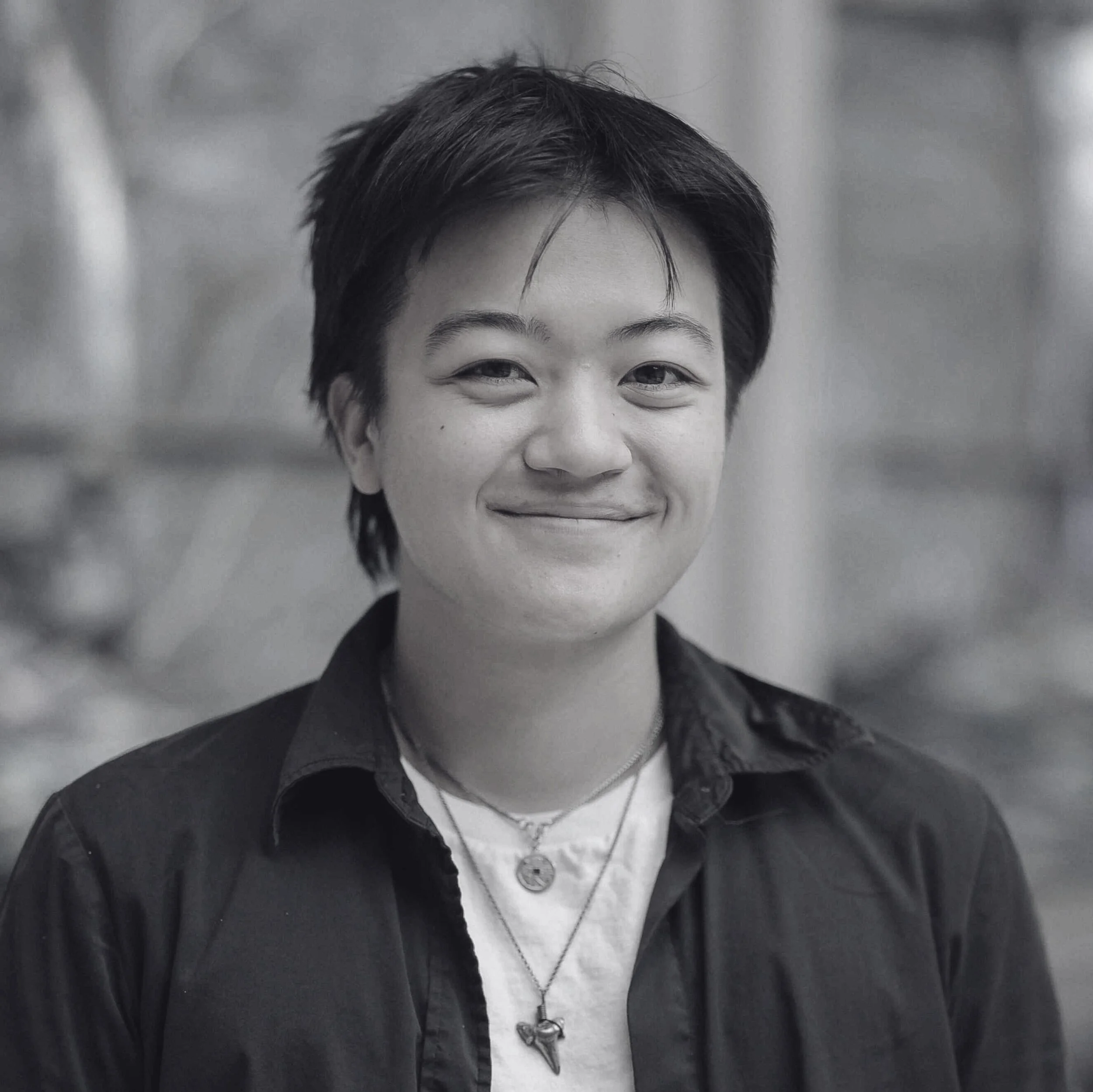Headshot of the poet from the shoulders up facing the camera with a closed-mouth smile. The poet has short dark hair and is wearing a white top with a dark overshirt, and a shark tooth on a chain around their neck.