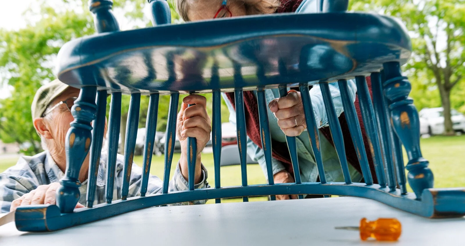 Two people work together outdoors to repair or tighten parts of a blue wooden chair, holding the spindles from underneath while a screwdriver rests on the table nearby.