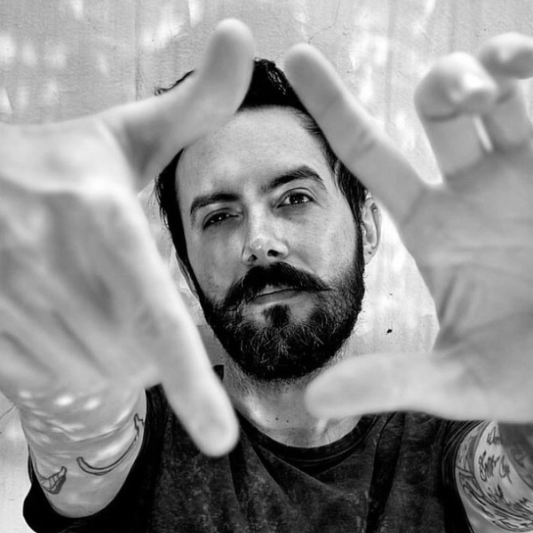 Black-and-white portrait of a bearded man holding his hands up to frame the camera, looking directly forward with a focused expression; tattoos are visible on his forearms.