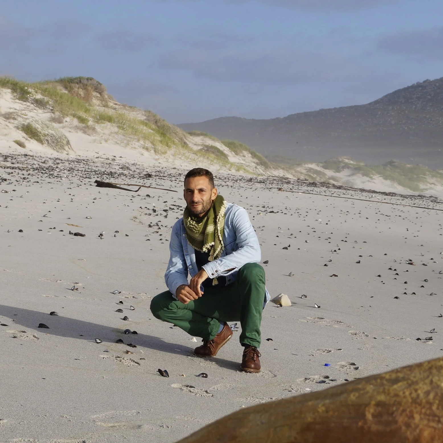Artist Vincenzo Cohen crouching on a sandy beach surrounded by dunes and rocky hills.