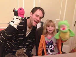 Piano teacher Andrew and his young student Clara posing with colorful puppets during a fun lesson.