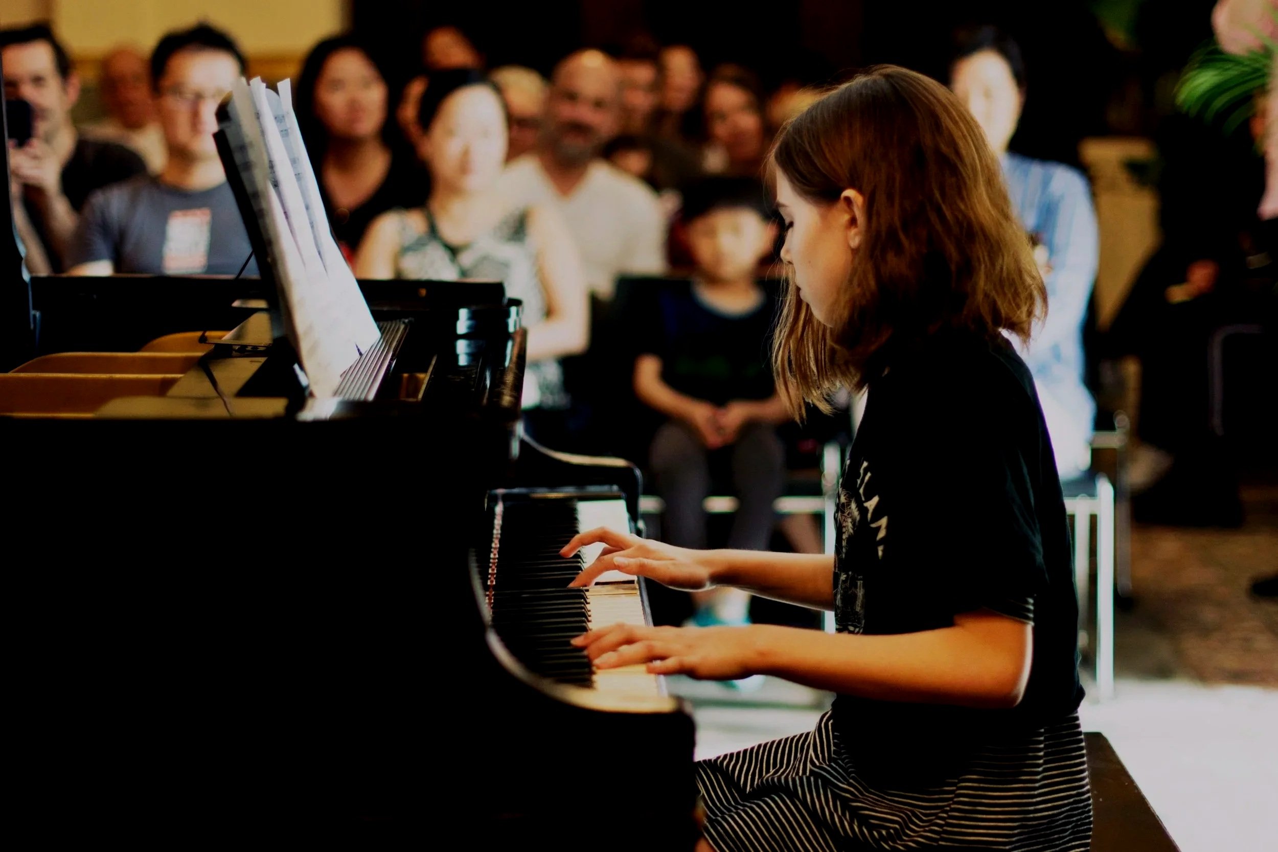 A young piano student performing at a recital, seated at a grand piano in front of an audience at A Piano Grows in Brooklyn.