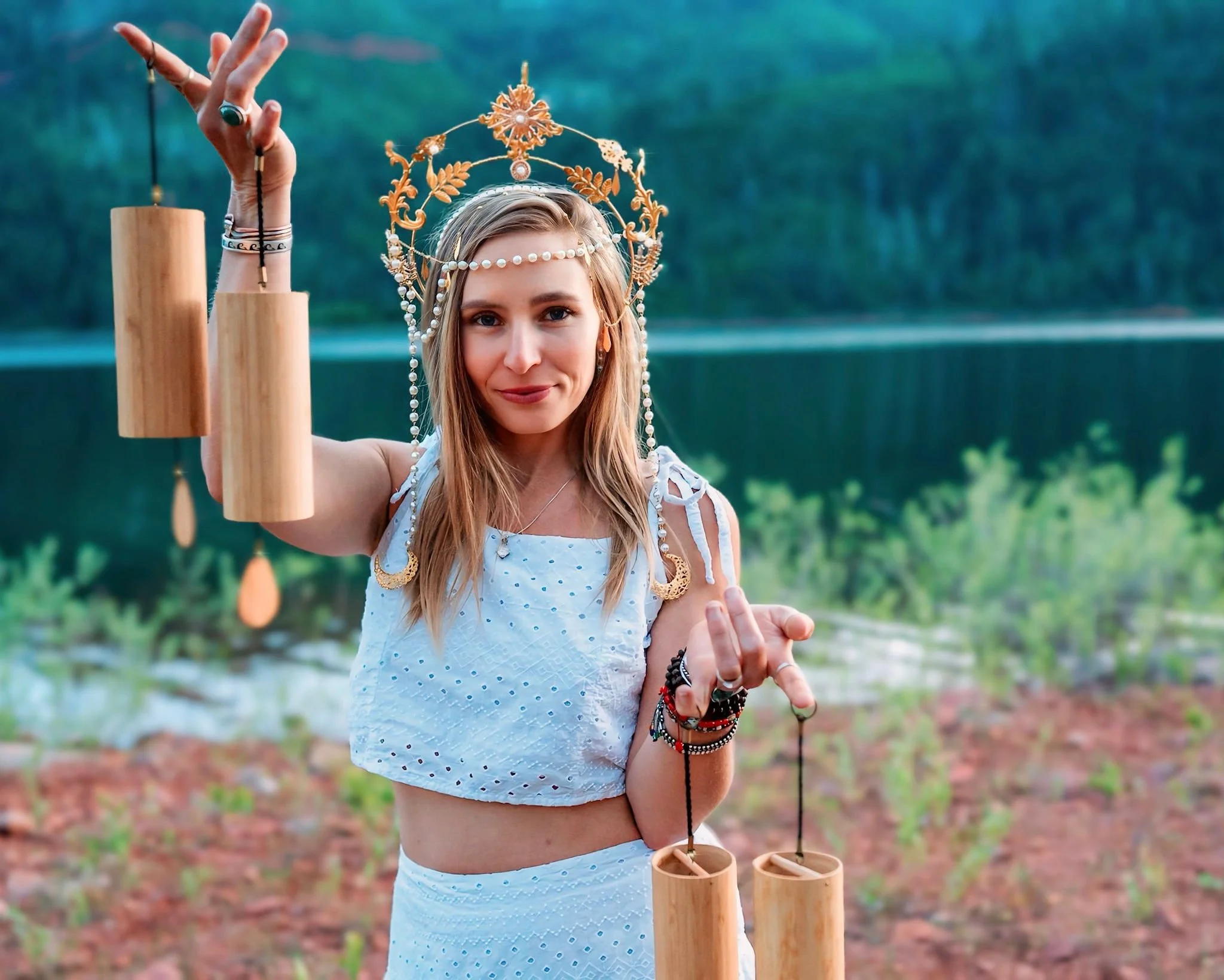 A woman wearing traditional or ceremonial attire with a decorative headpiece, holding wooden wind chimes, standing outdoors near a body of water with trees in the background.