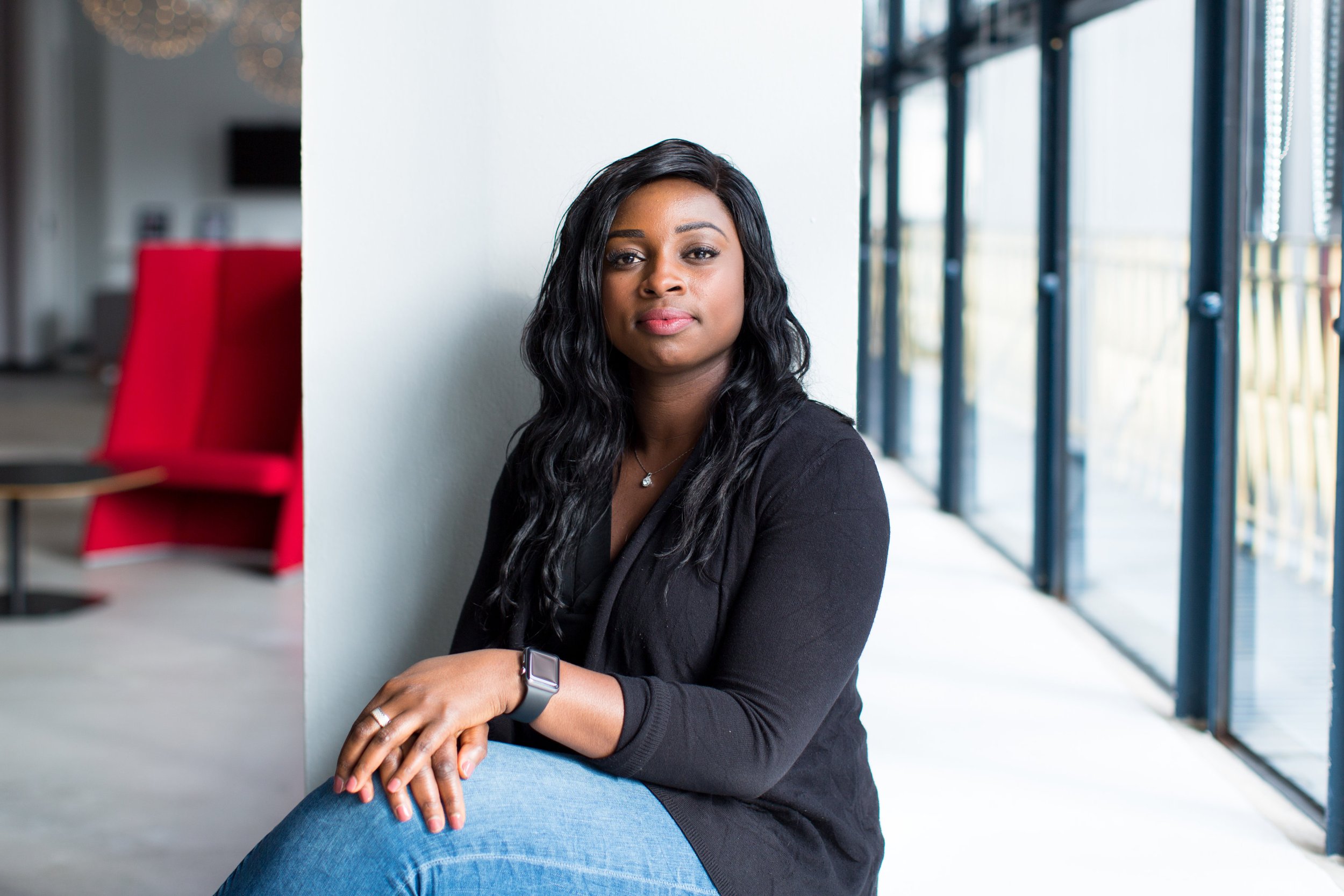 A woman with long black hair wearing a black top, blue jeans, and a smartwatch sitting by a large window in a modern indoor space.