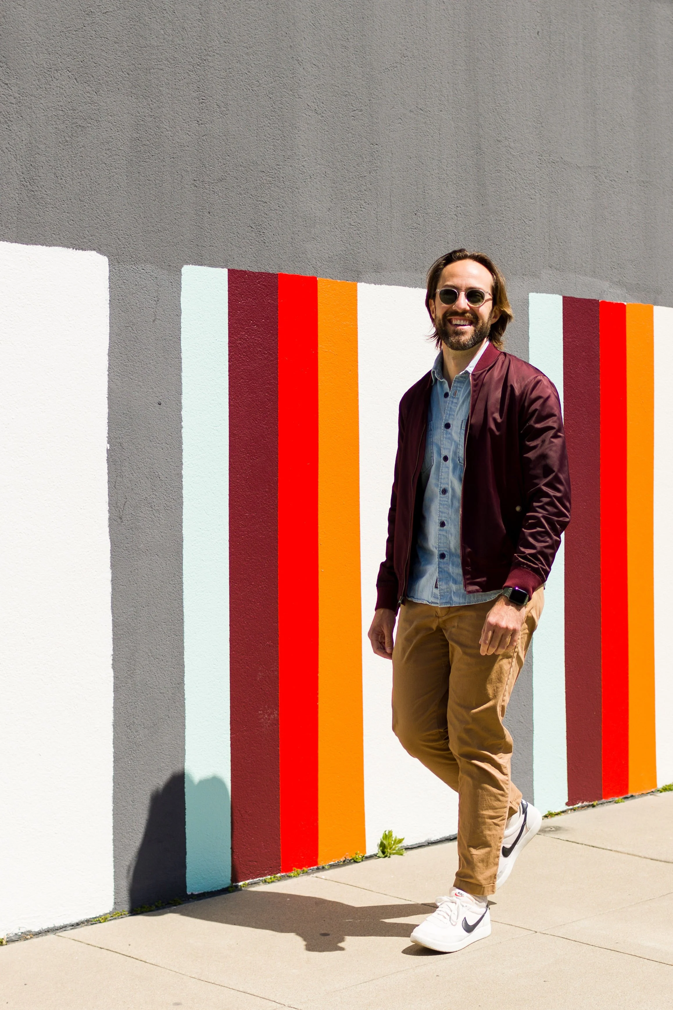 A man with sunglasses, a beard, wearing a maroon jacket, light blue shirt, khaki pants, and white sneakers, standing against a colorful wall with vertical stripes in shades of red, orange, and light blue.