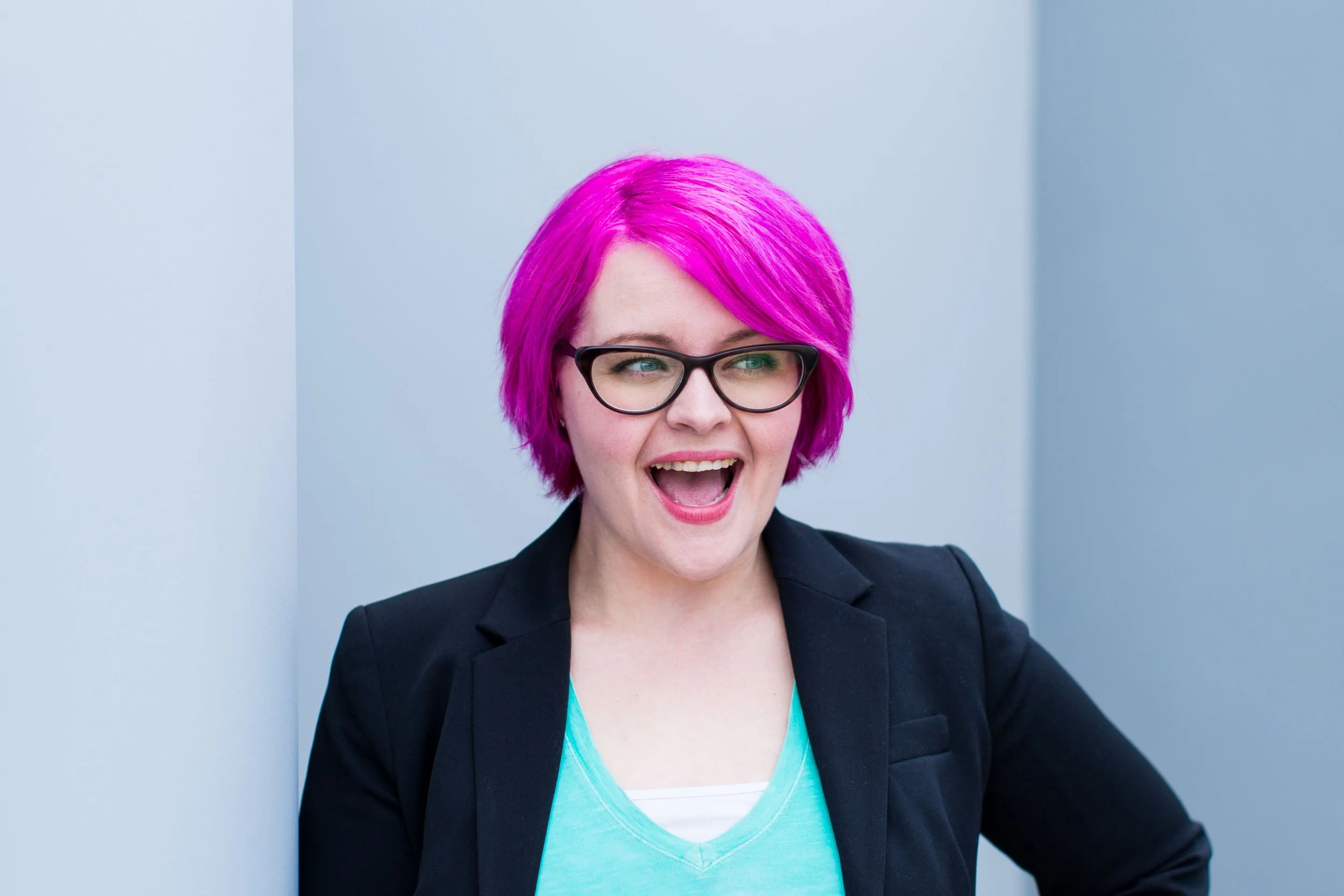 A woman with bright pink hair, black glasses, wearing a light blue shirt and black blazer, smiling and looking to her left.