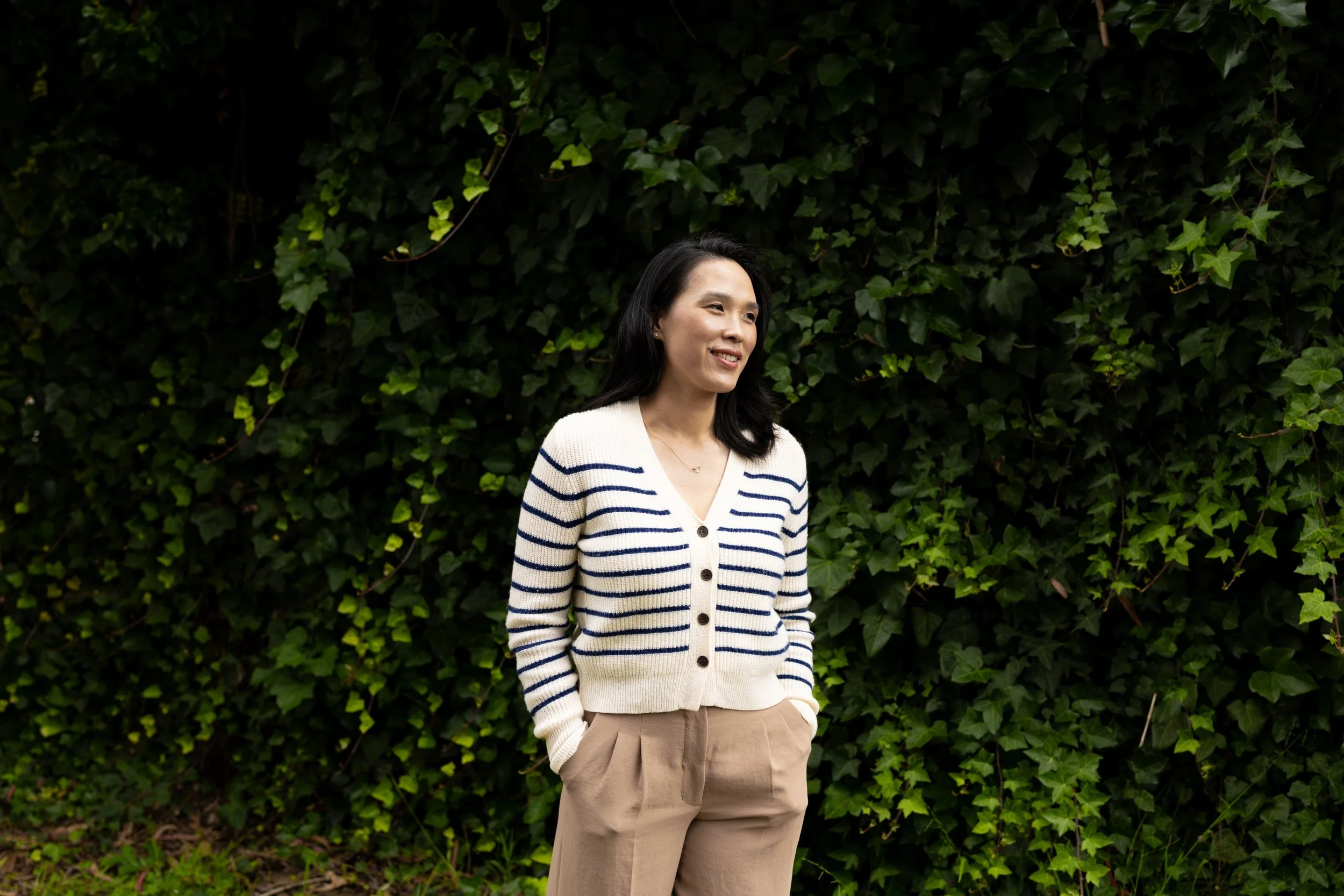 A woman standing outdoors in front of a dense green ivy wall, smiling and looking to her left. She is wearing a cream-colored cardigan with navy blue stripes and beige pants.