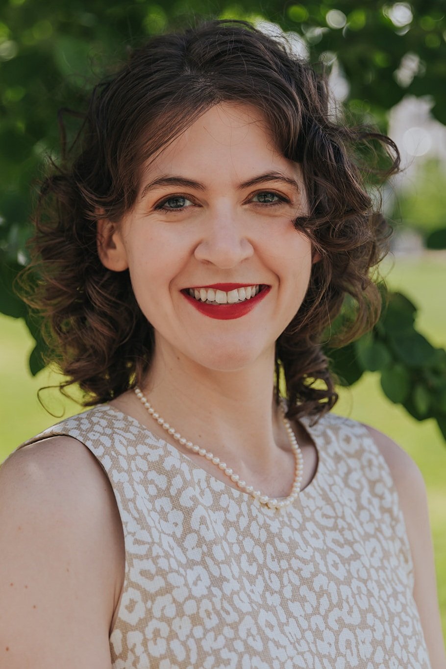 A white woman with curly brown hair and red lipstick. She is smiling at the camera.