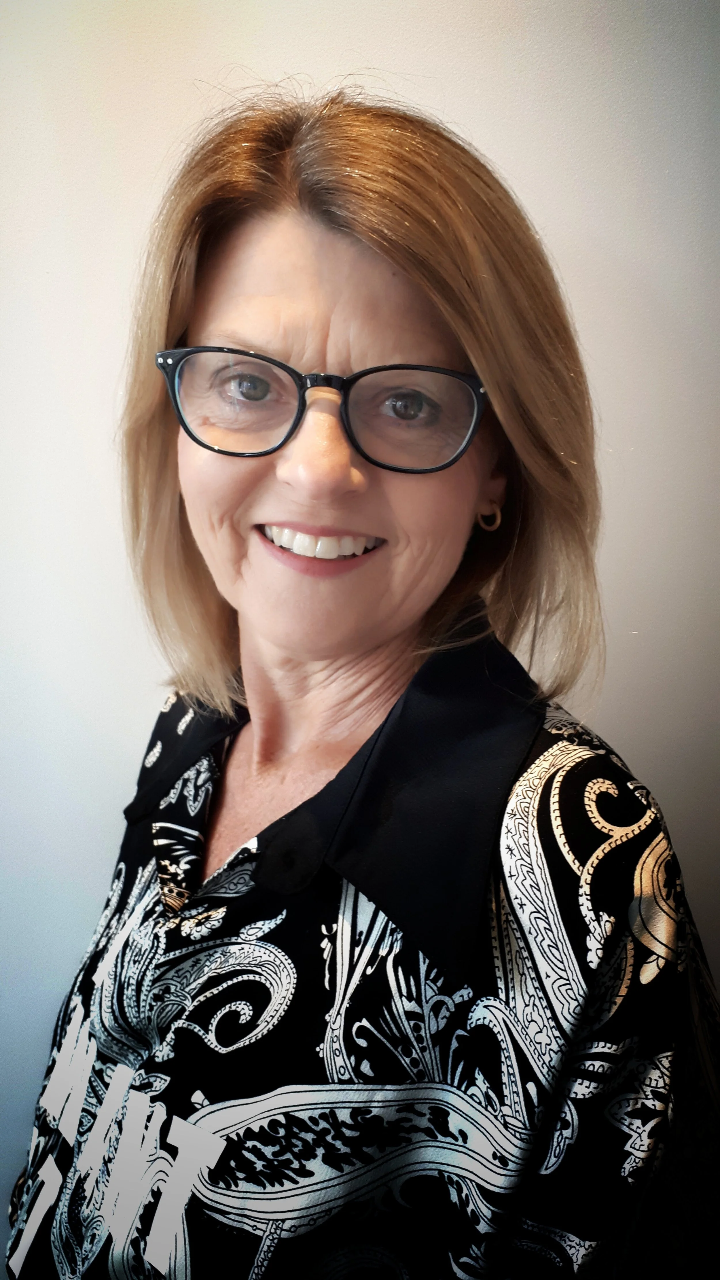 Woman with glasses smiling, wearing a patterned black and white blouse, standing against a plain background.