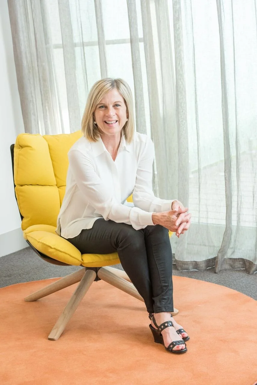 Woman sitting in a yellow chair with a relaxed posture, wearing a white blouse and black pants, against a backdrop of sheer curtains.