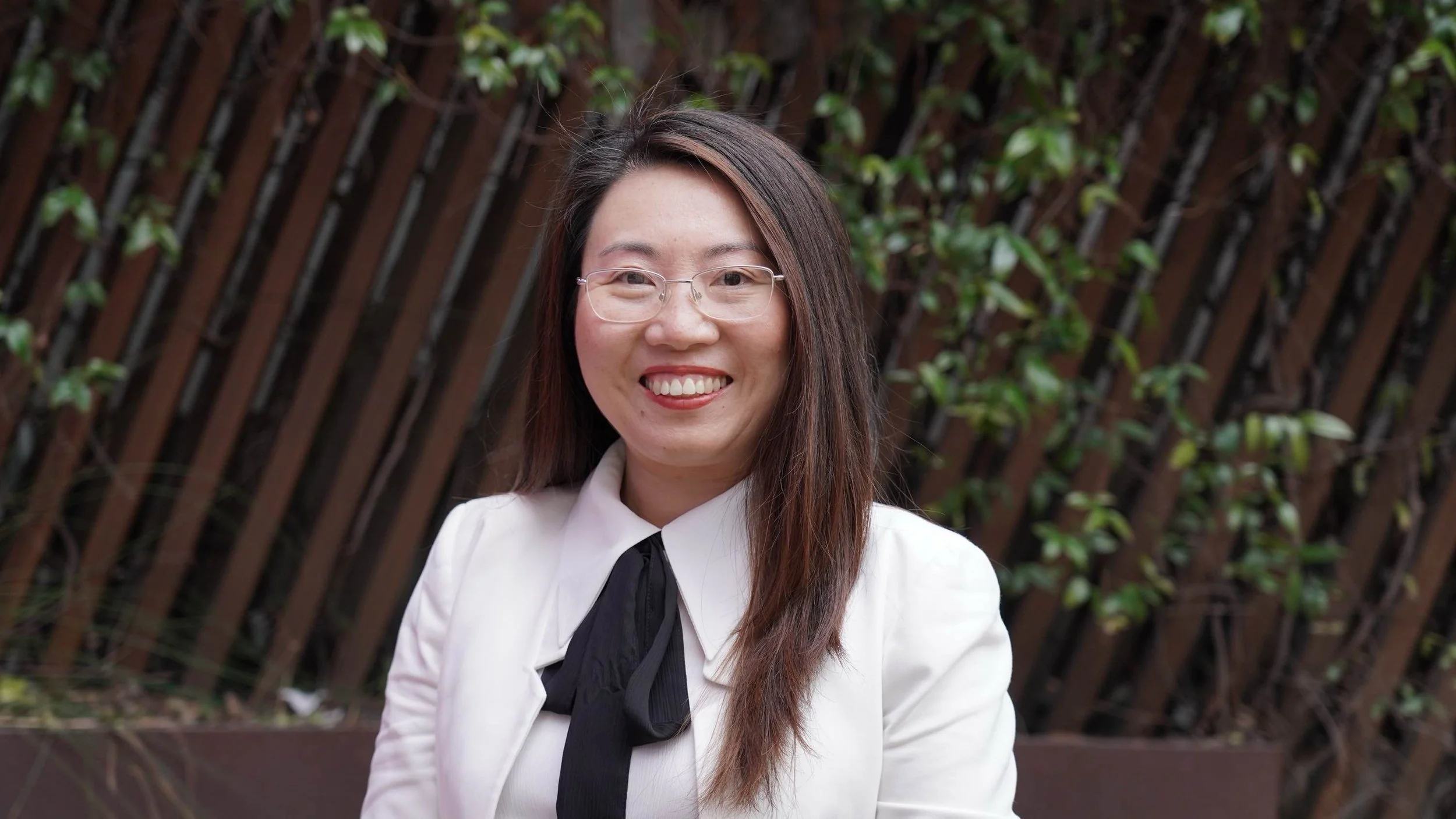 Smiling person in glasses and formal attire with greenery and wooden fence in background.