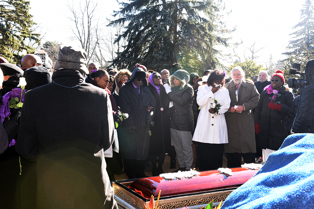 A grieving Olivia Grange in purple scarf and Gifford Walker in purple shirt with black hat at the burial site