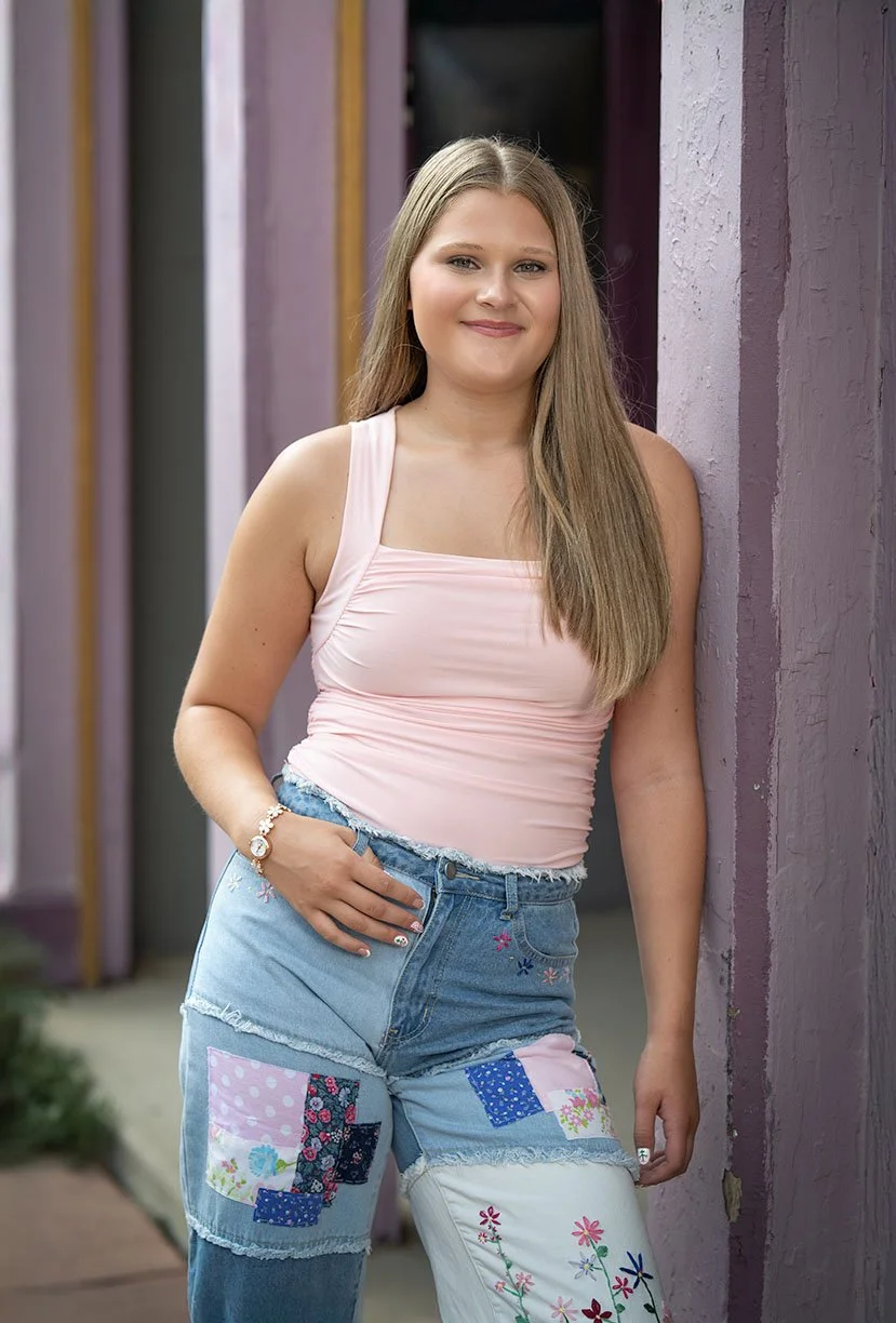 Young woman with long light brown hair smiling and standing outdoors against purple painted walls, wearing a pink sleeveless top and multicolored patchwork jeans.