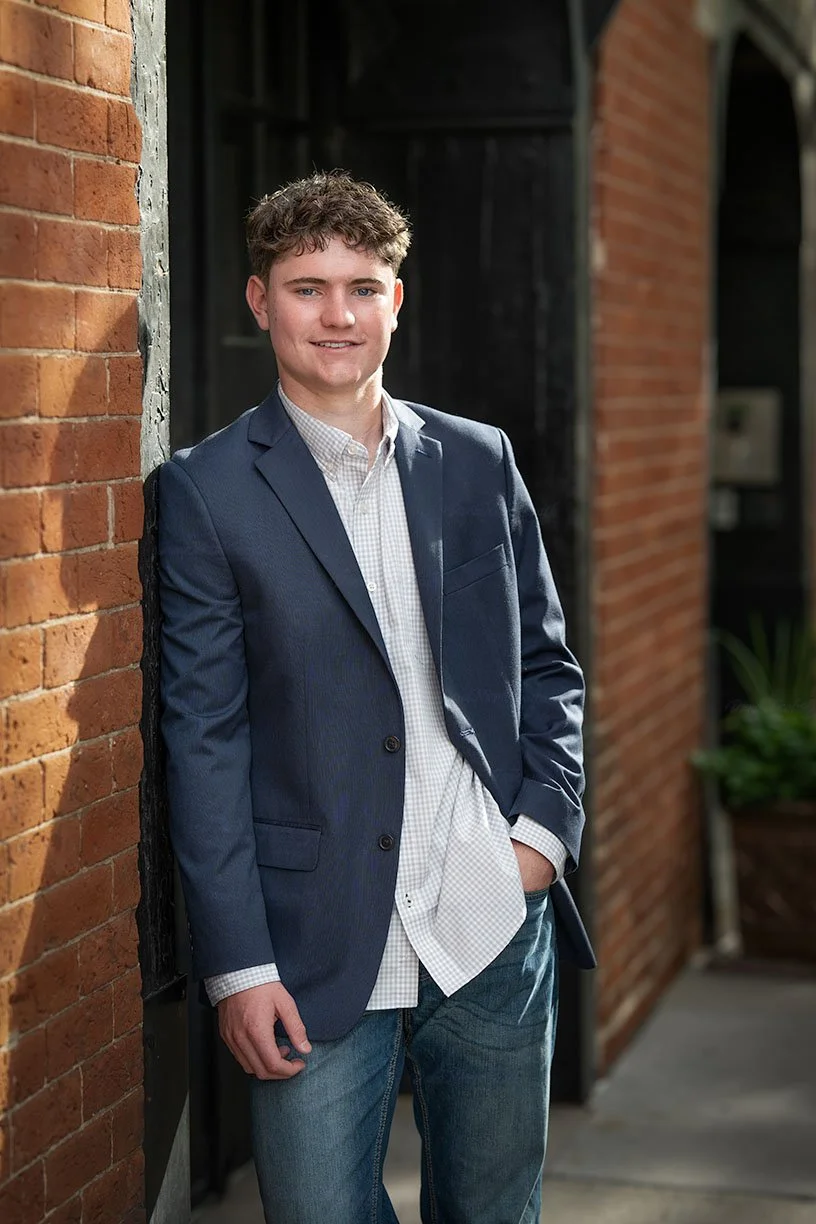 Young man in a navy blazer and light-colored shirt standing against a brick wall, smiling.